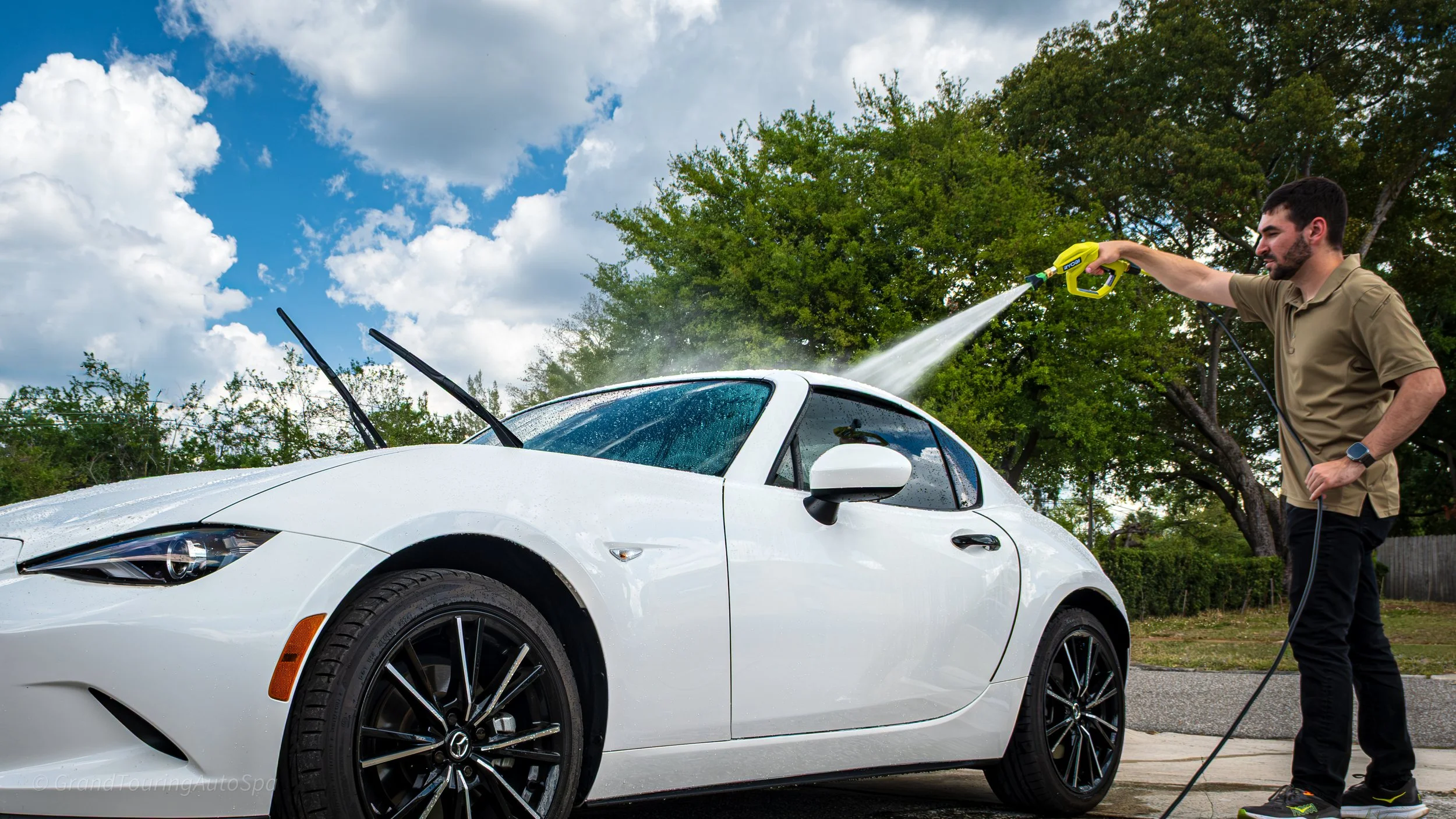 A man washing a white sports car with a hose outdoors under a partly cloudy sky, with green trees in the background.