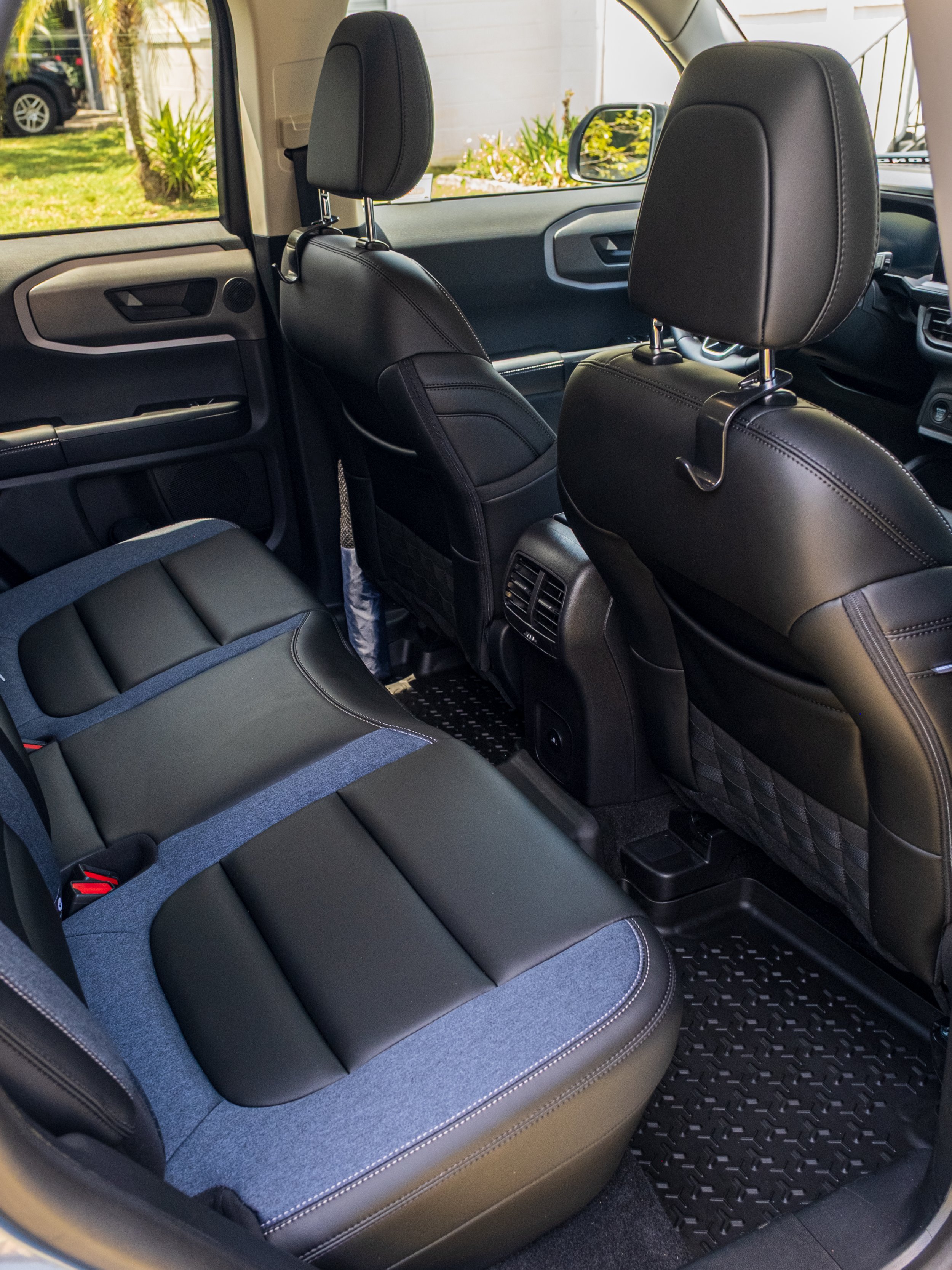 The interior of a vehicle showing the back seat and front seats with black and gray upholstery, a dashboard, and a view of the outside with houses and greenery.