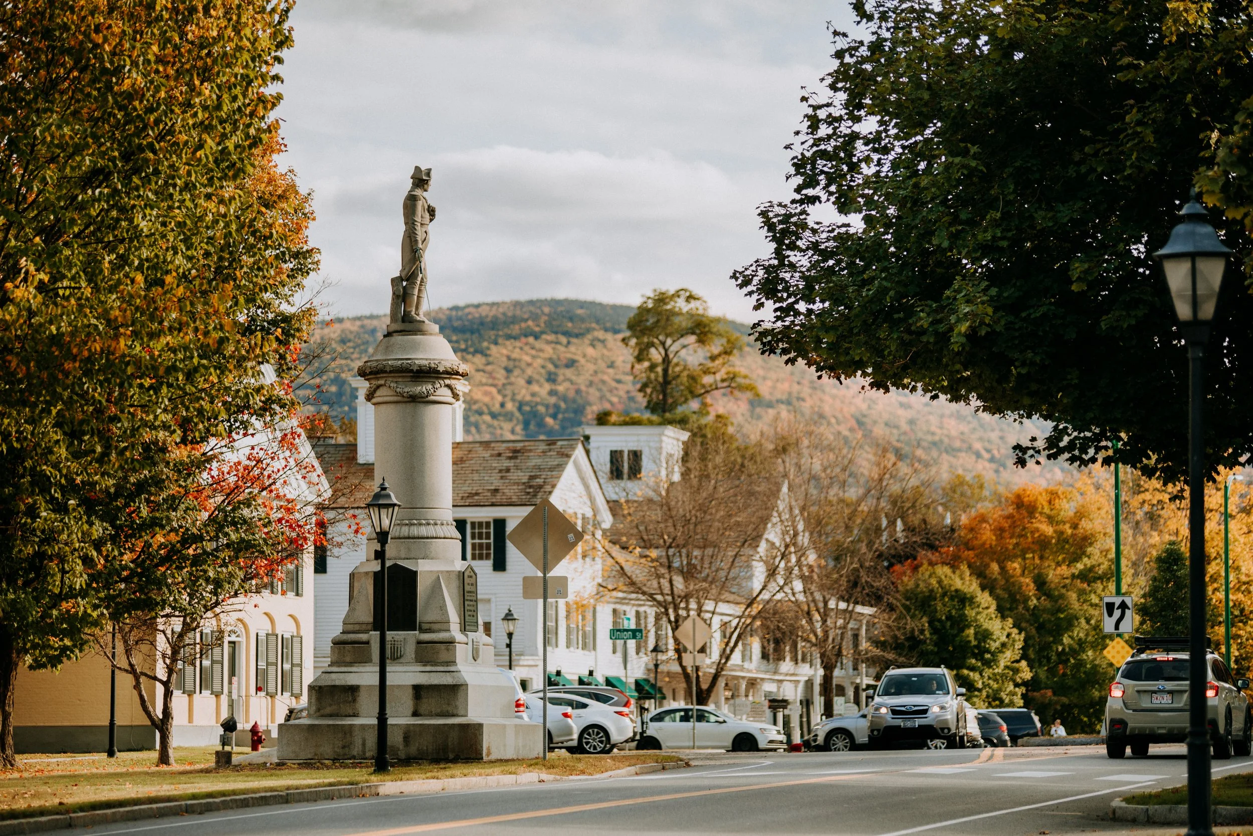 A street scene with a monument of a historical figure on a pedestal, surrounded by trees with fall foliage, parked cars, and white buildings in the background, hills in the distance.