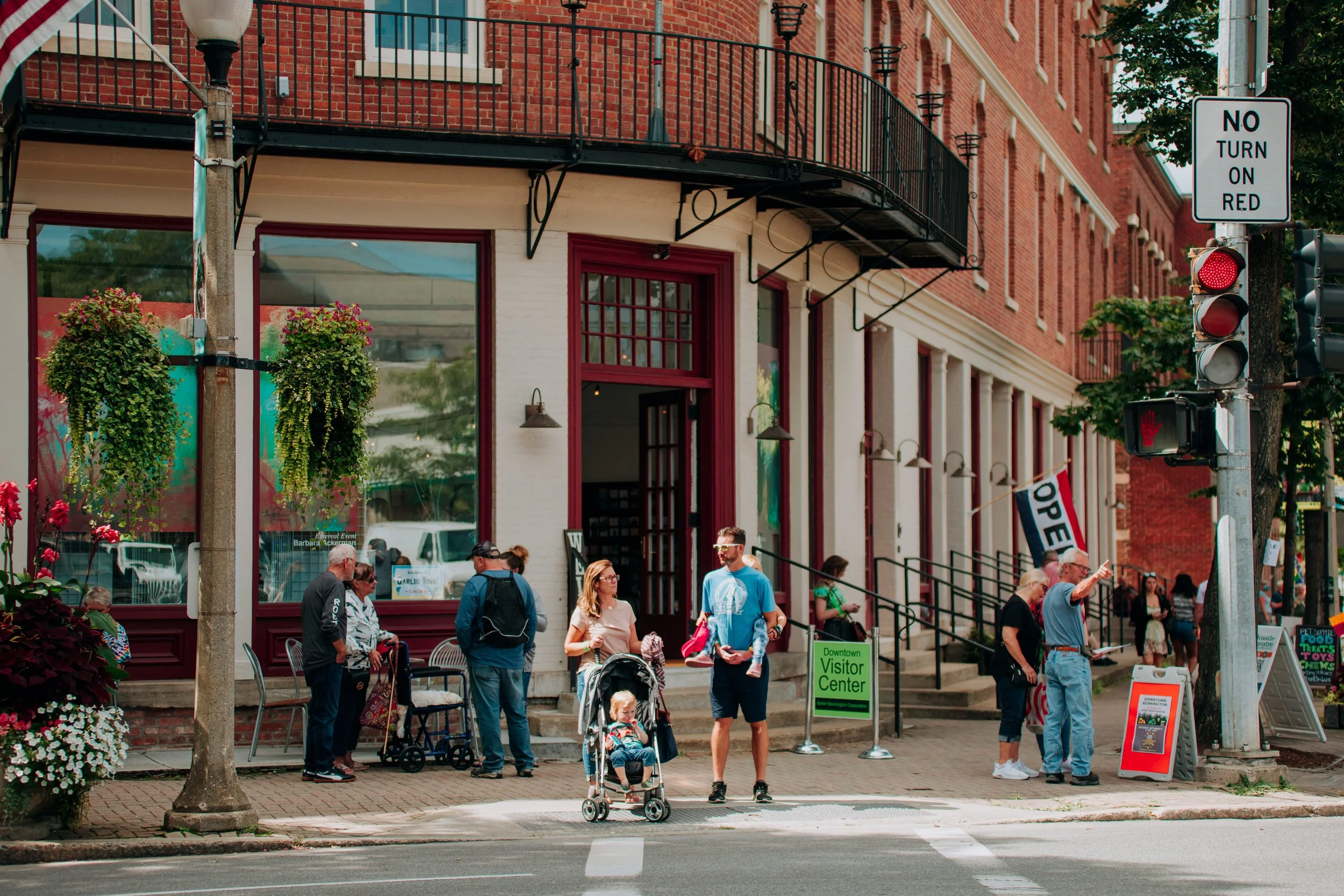 People waiting at a crosswalk in front of a brick building with an open sign, a traffic light, and a 'No Turn on Red' sign in an urban area.