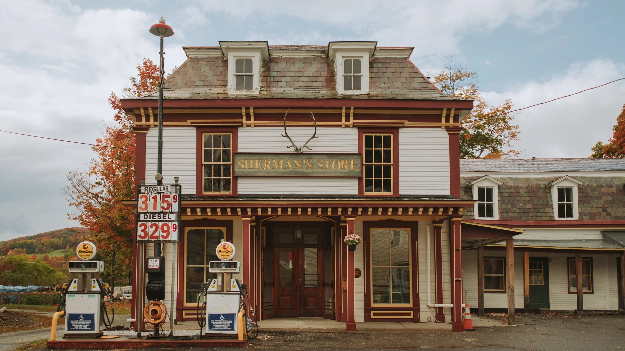 A vintage two-story store named Sherman’s Store with gas pumps in front and autumn trees in the background.