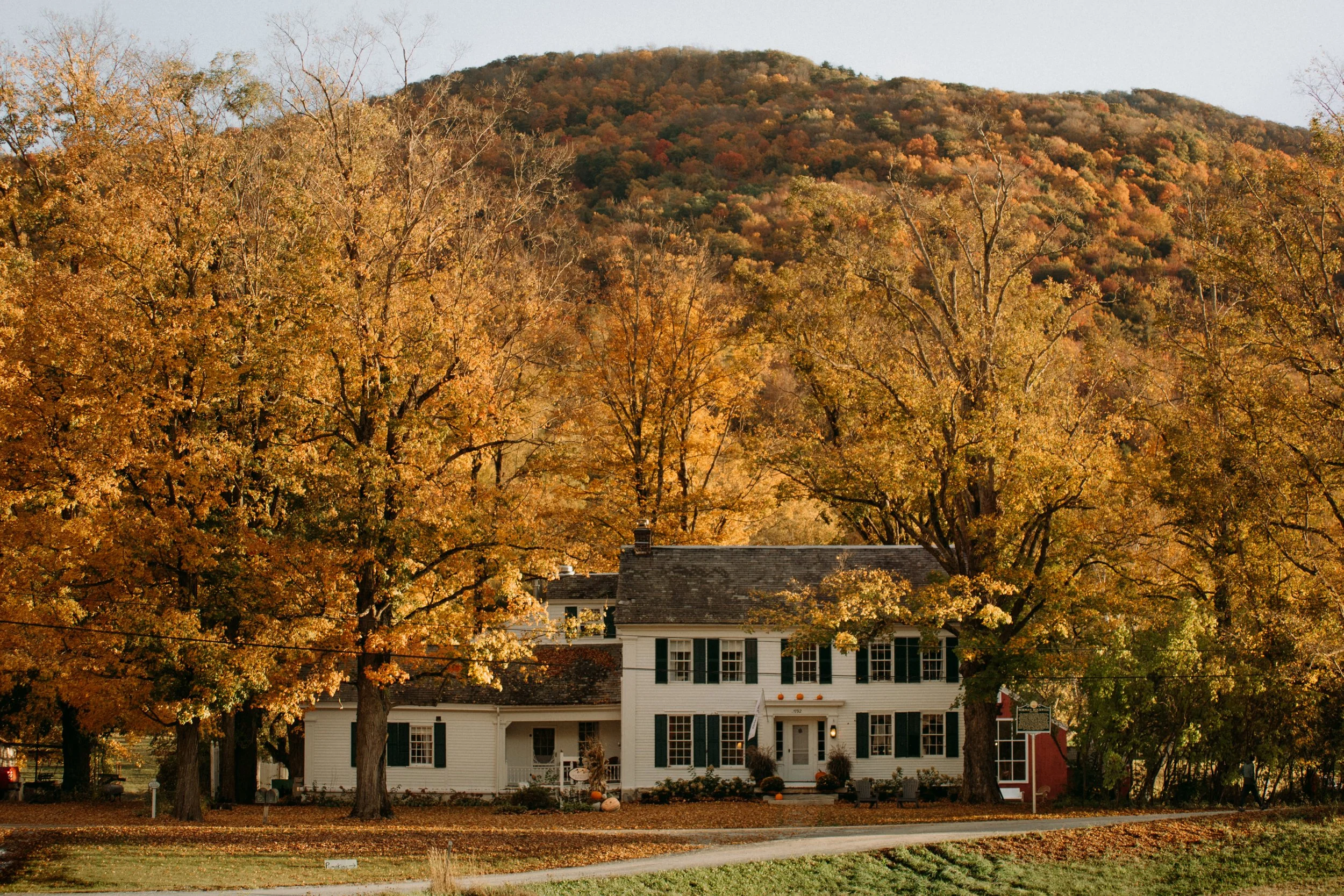 A large white house with black shutters, surrounded by fall foliage and large trees in autumn colors. The house is located in front of a wooded hill with similarly colored trees.