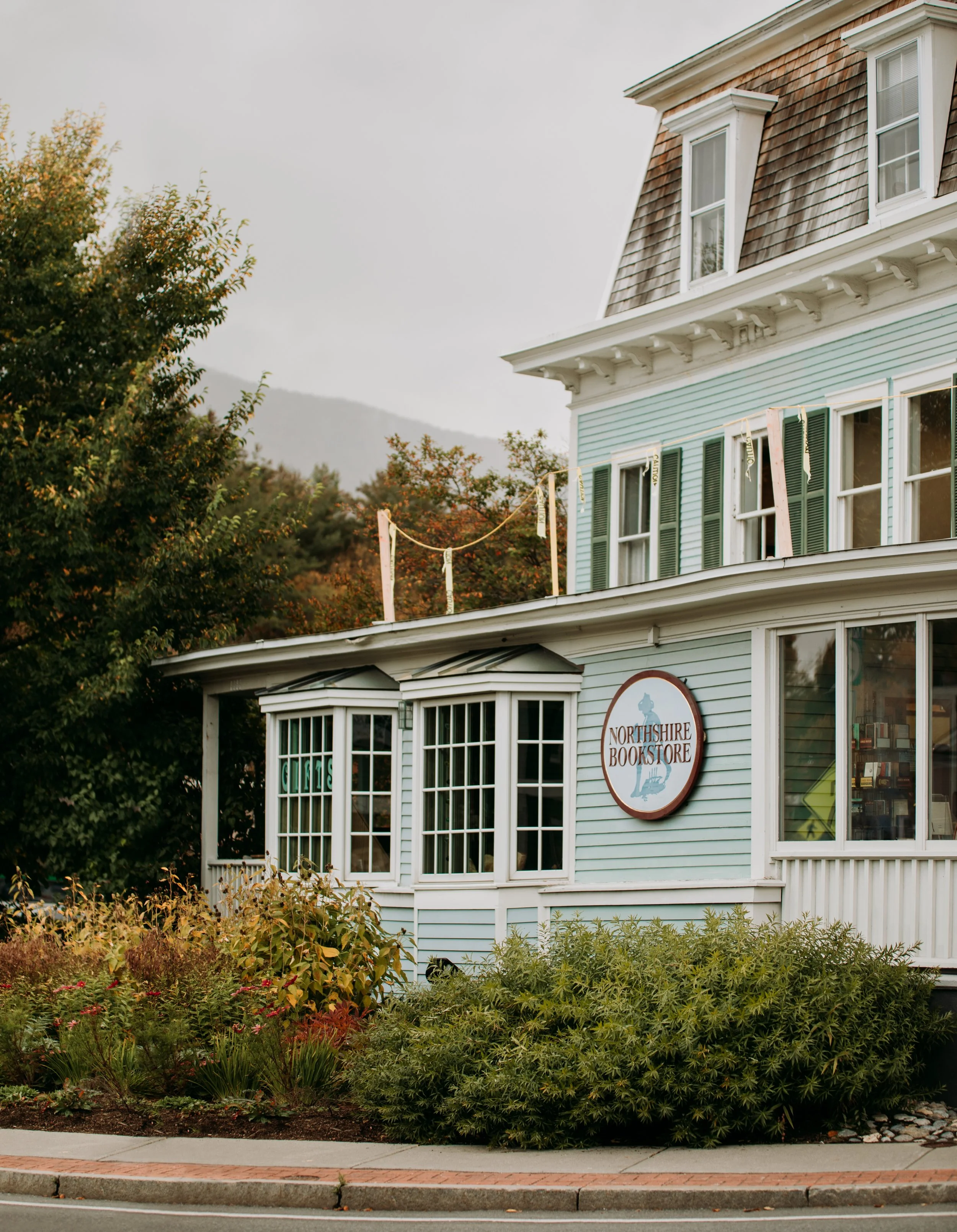 Exterior of a light blue building with white trim and a round sign that reads 'Northshire Bookstore', surrounded by lush green bushes and trees, with mountains in the background.