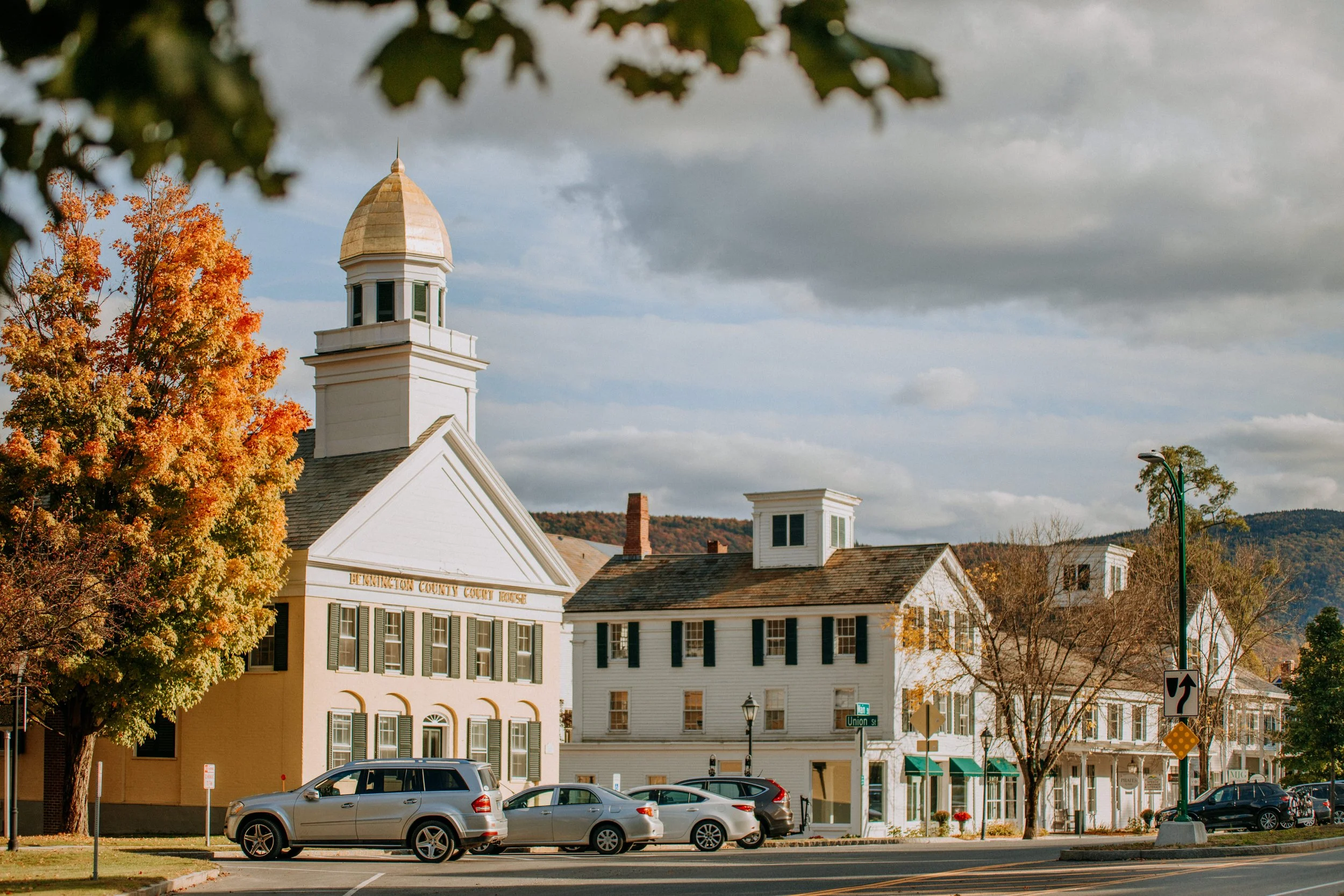 A small town street with a courthouse, primarily white with black shutters, and several nearby buildings with green awnings. Cars are parked along the street, and trees with fall foliage are visible.