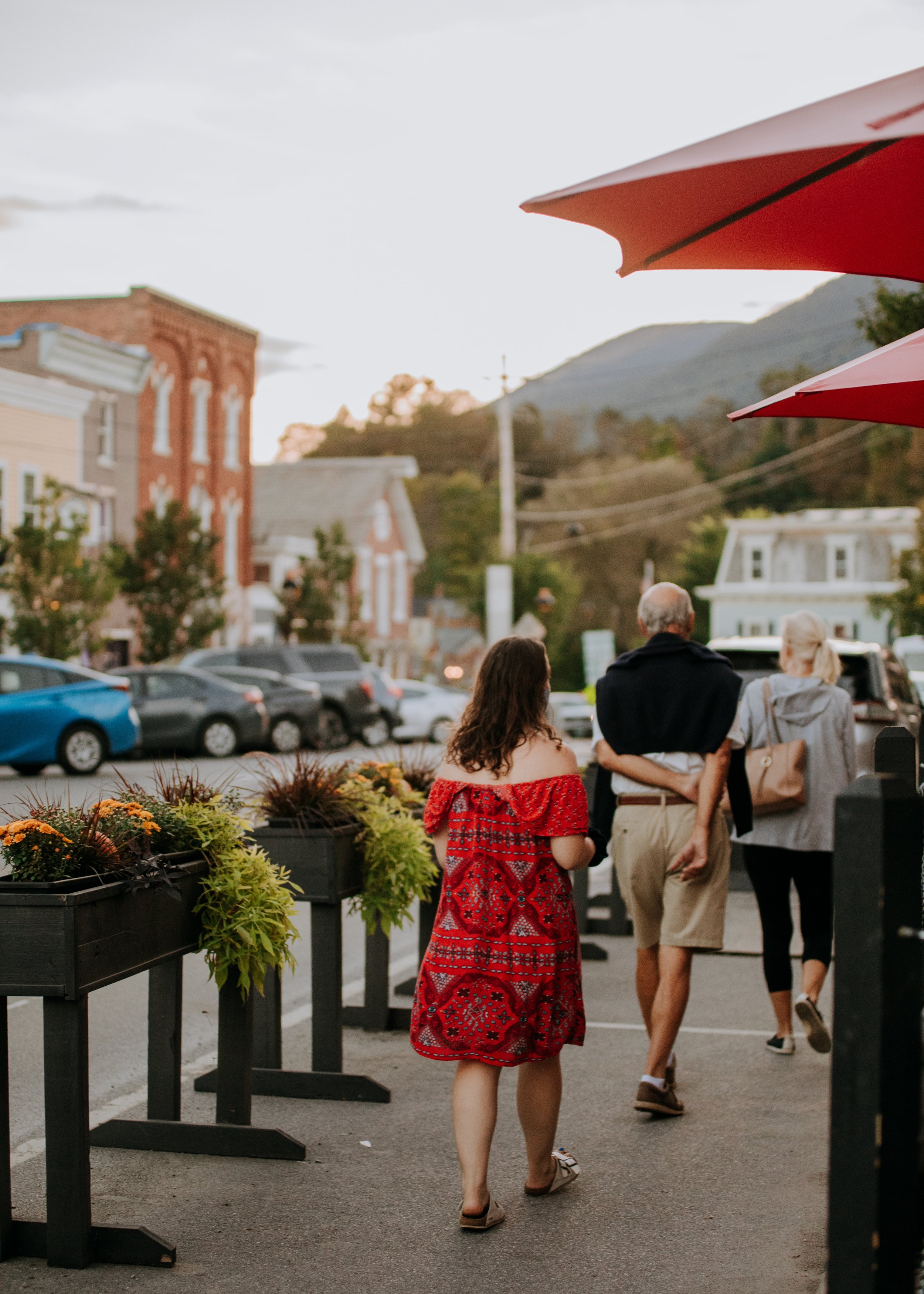 People walking down a sidewalk in a small town, with parked cars, historic buildings, and a mountain in the background, during sunset.