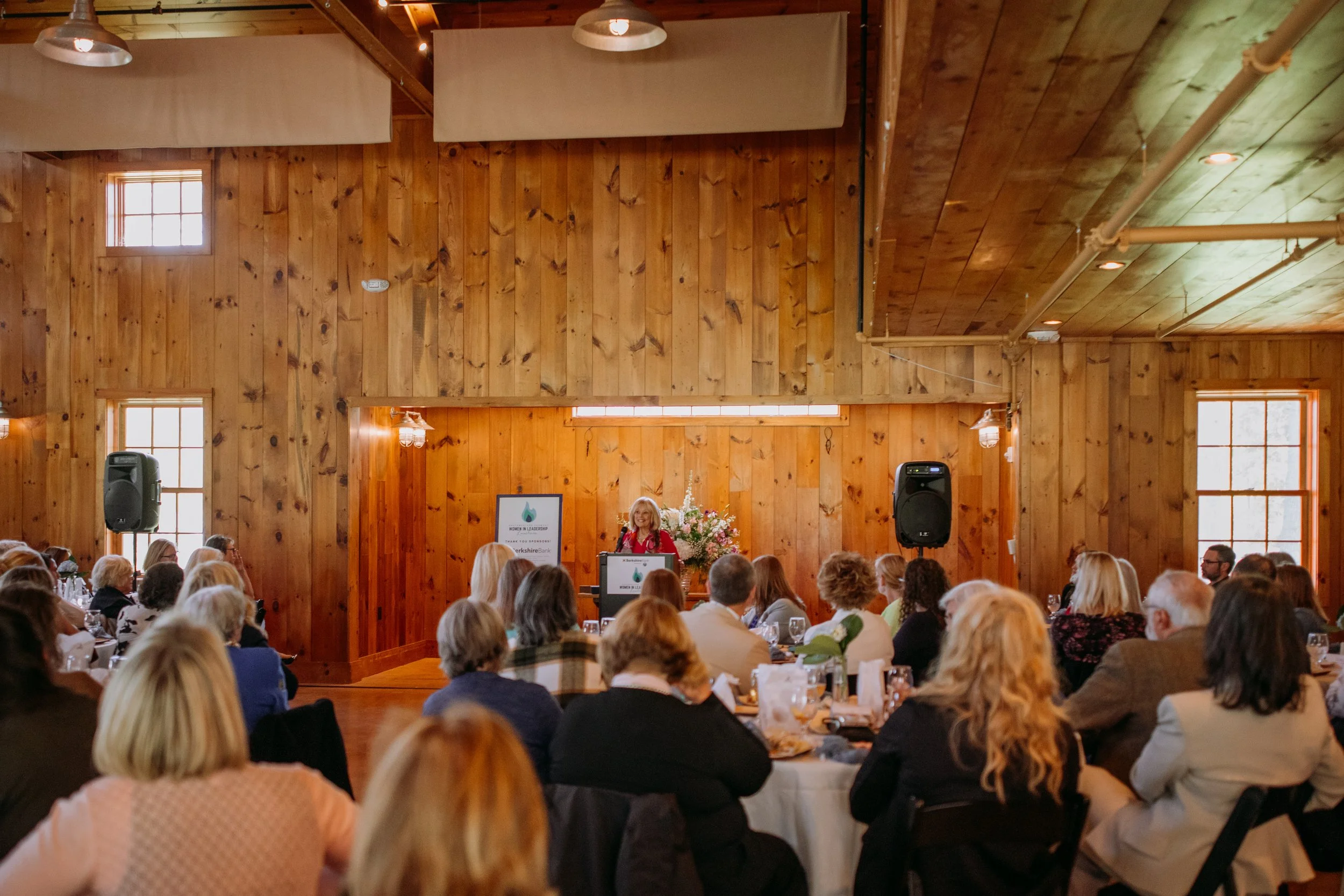 A woman speaks at a podium during a conference or event in a wooden hall with seated attendees listening.