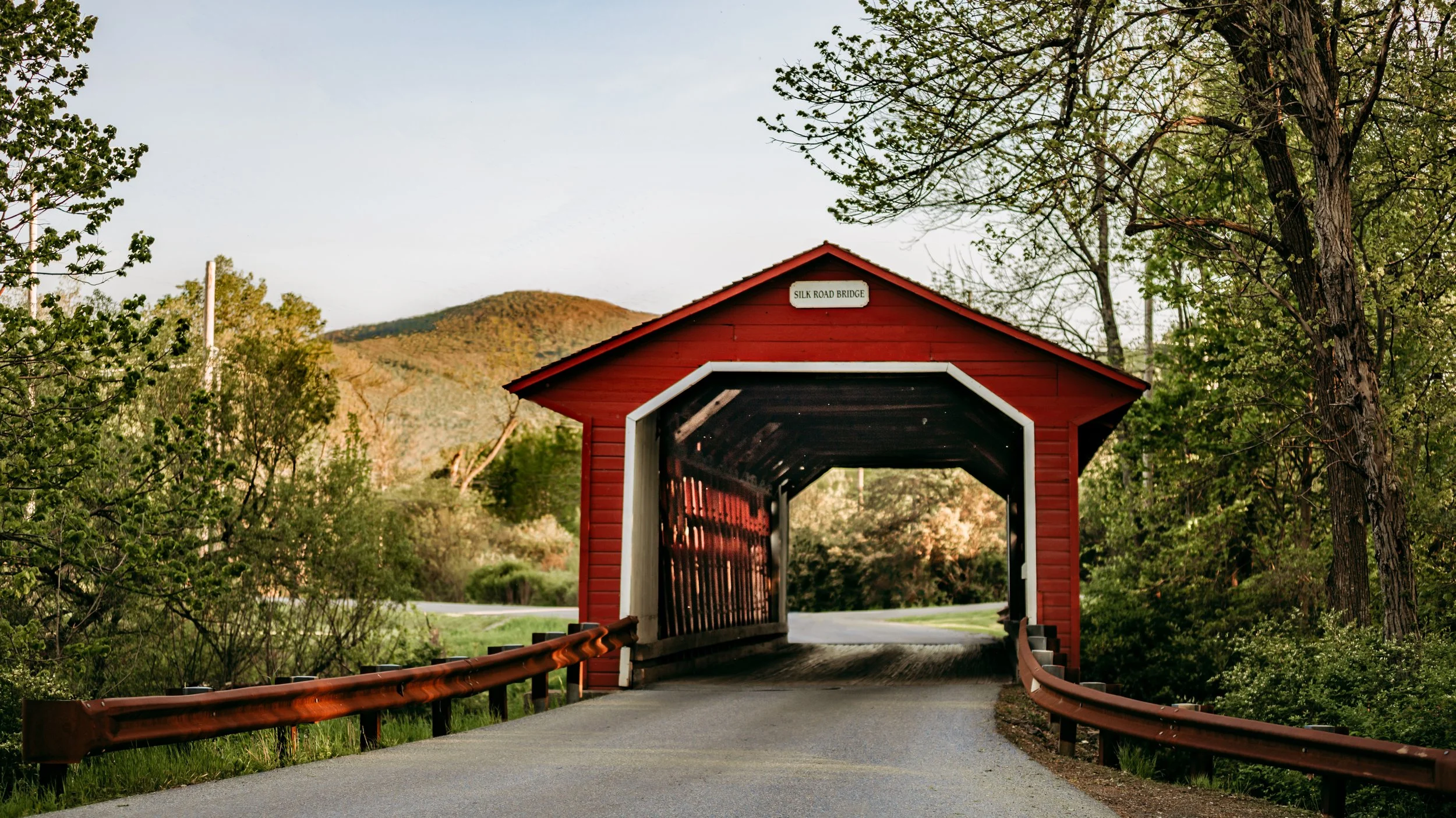 Red covered bridge with white trim over a narrow road surrounded by green trees and hills in the background.