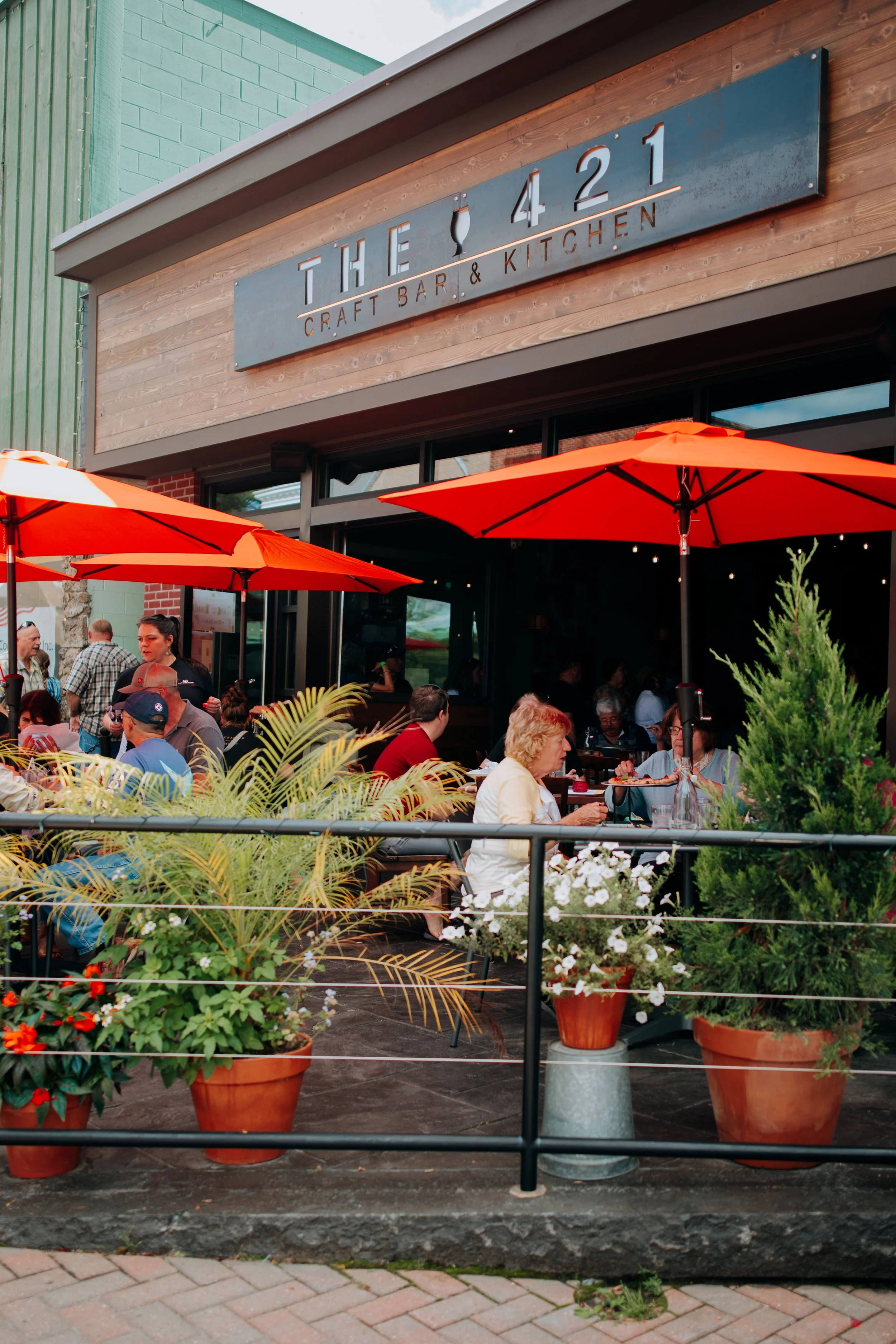 Outdoor patio of a craft bar and kitchen with people dining under orange umbrellas, potted plants, and a sign that reads 'The 421: Craft Bar & Kitchen'.