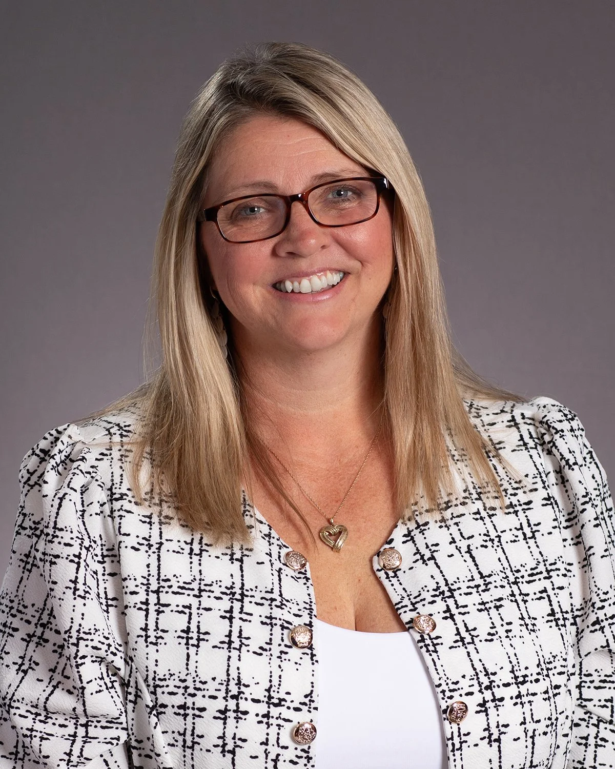 A professional woman with blonde hair, glasses, and hoop earrings, smiling in a studio portrait against a gray background.