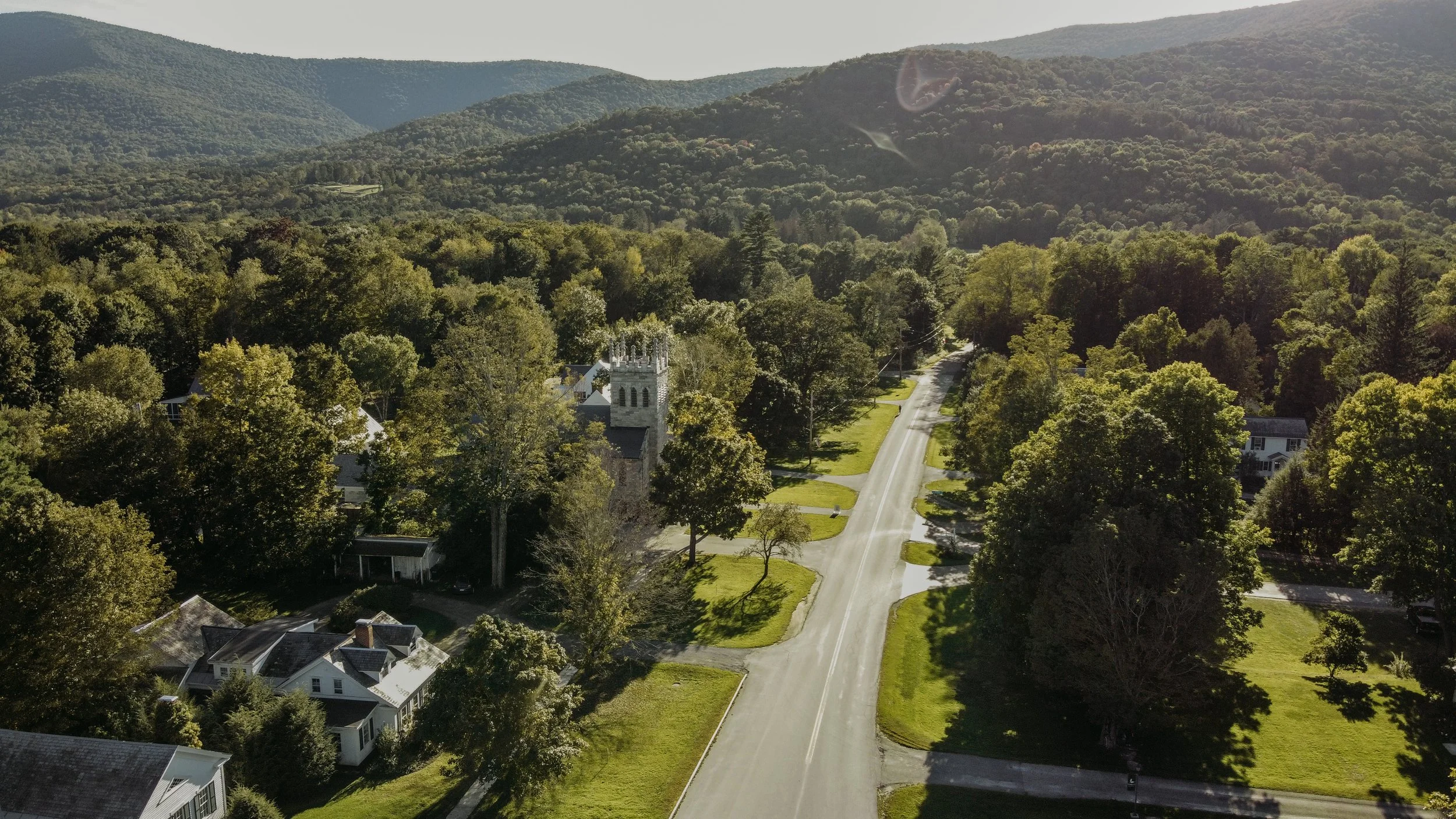 Aerial view of a small town street lined with trees and houses, surrounded by green wooded hills and mountains in the background, with the sun casting shadows from the trees.