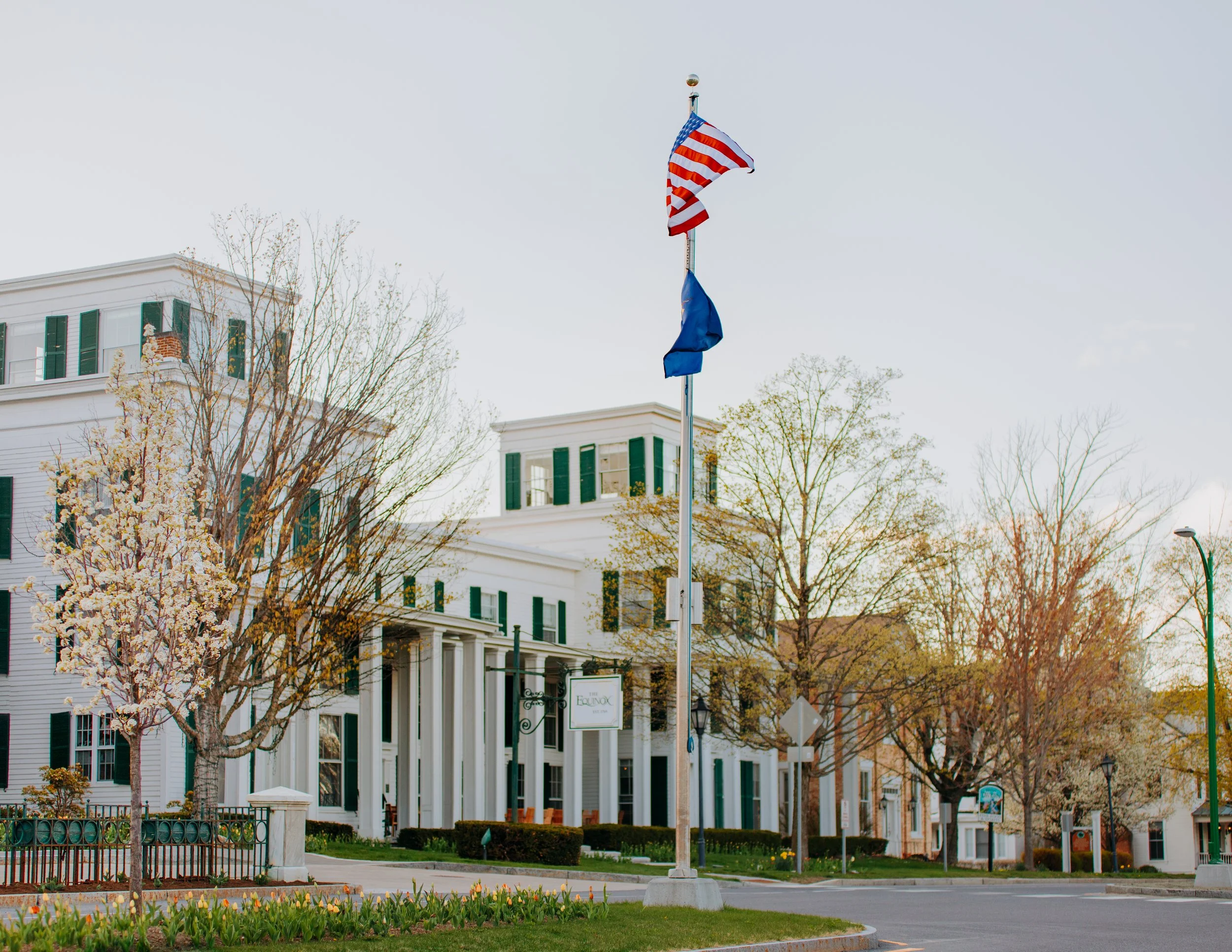 Street view of a white building with a sign reading "Eden Roc" surrounded by trees, some with blooming flowers, and a flagpole flying the American flag and another blue flag in a small town or village.