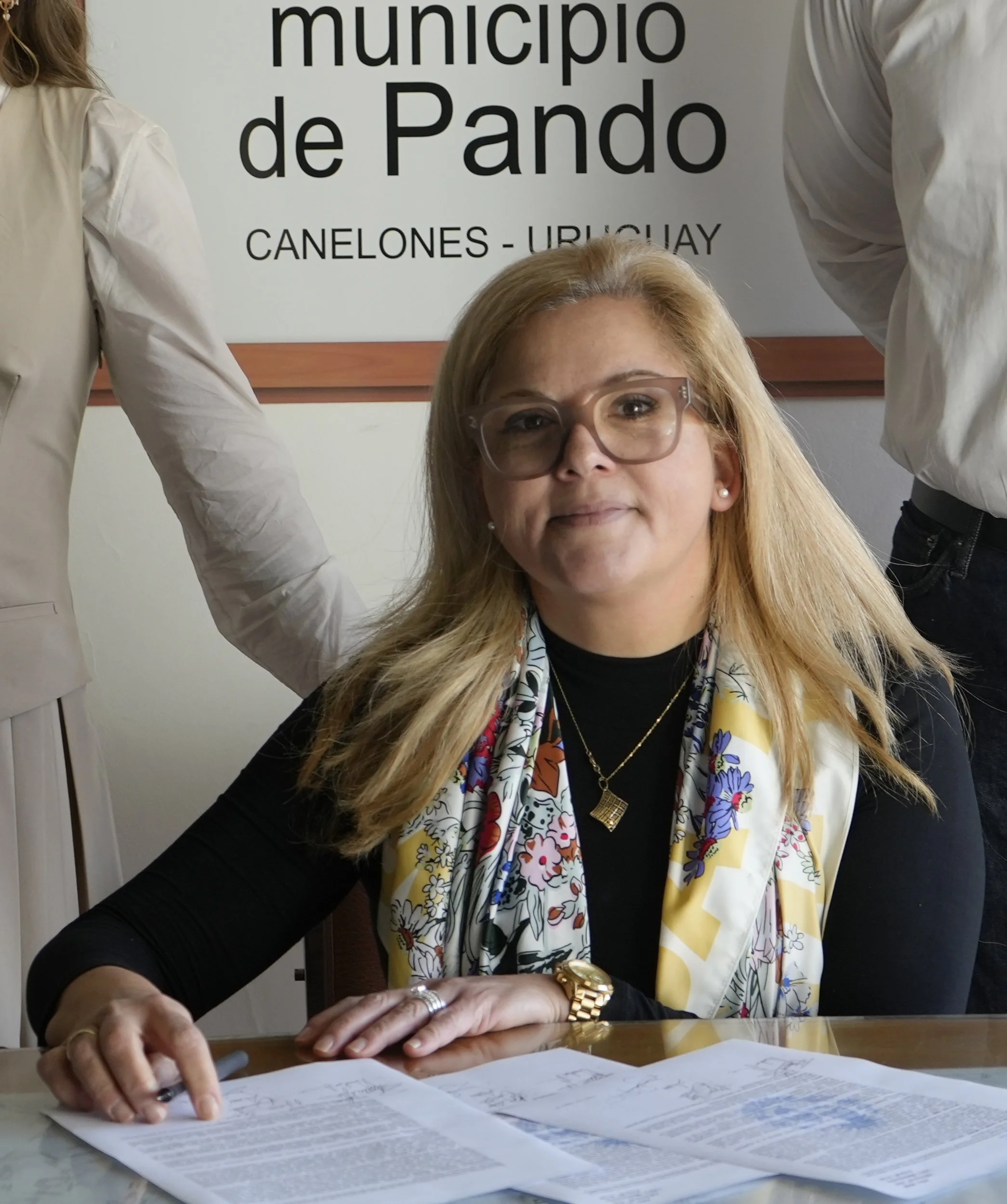 Una mujer con gafas y accesorios dorados, sentada en una mesa con documentos, en una oficina del municipio de Pando, Canelones, Uruguay, con una señal en la pared.