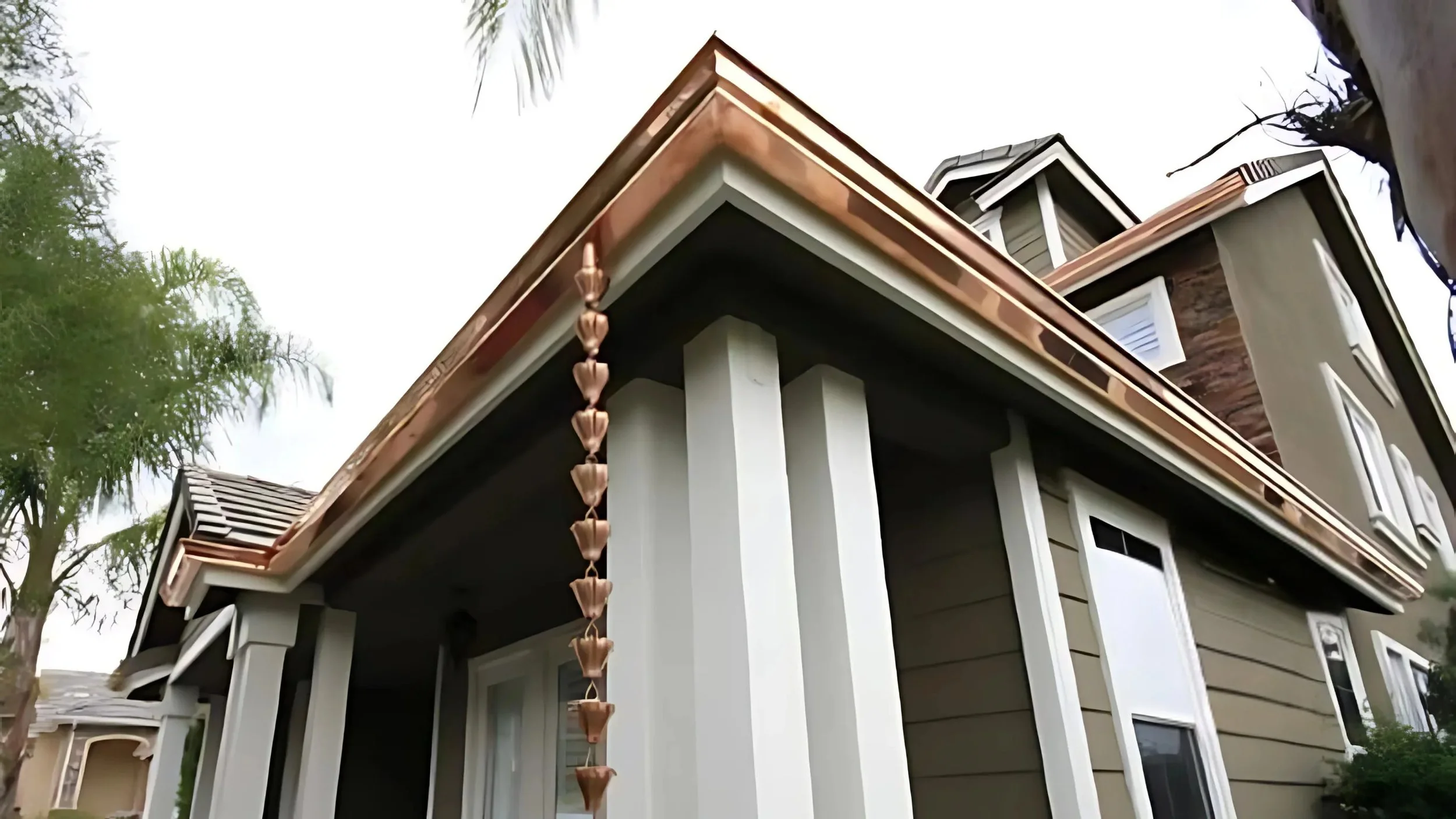 Close-up view of a two-story house with beige siding, white trim, copper gutters, and a decorative hanging planter on the corner of the roof, with trees in the background.