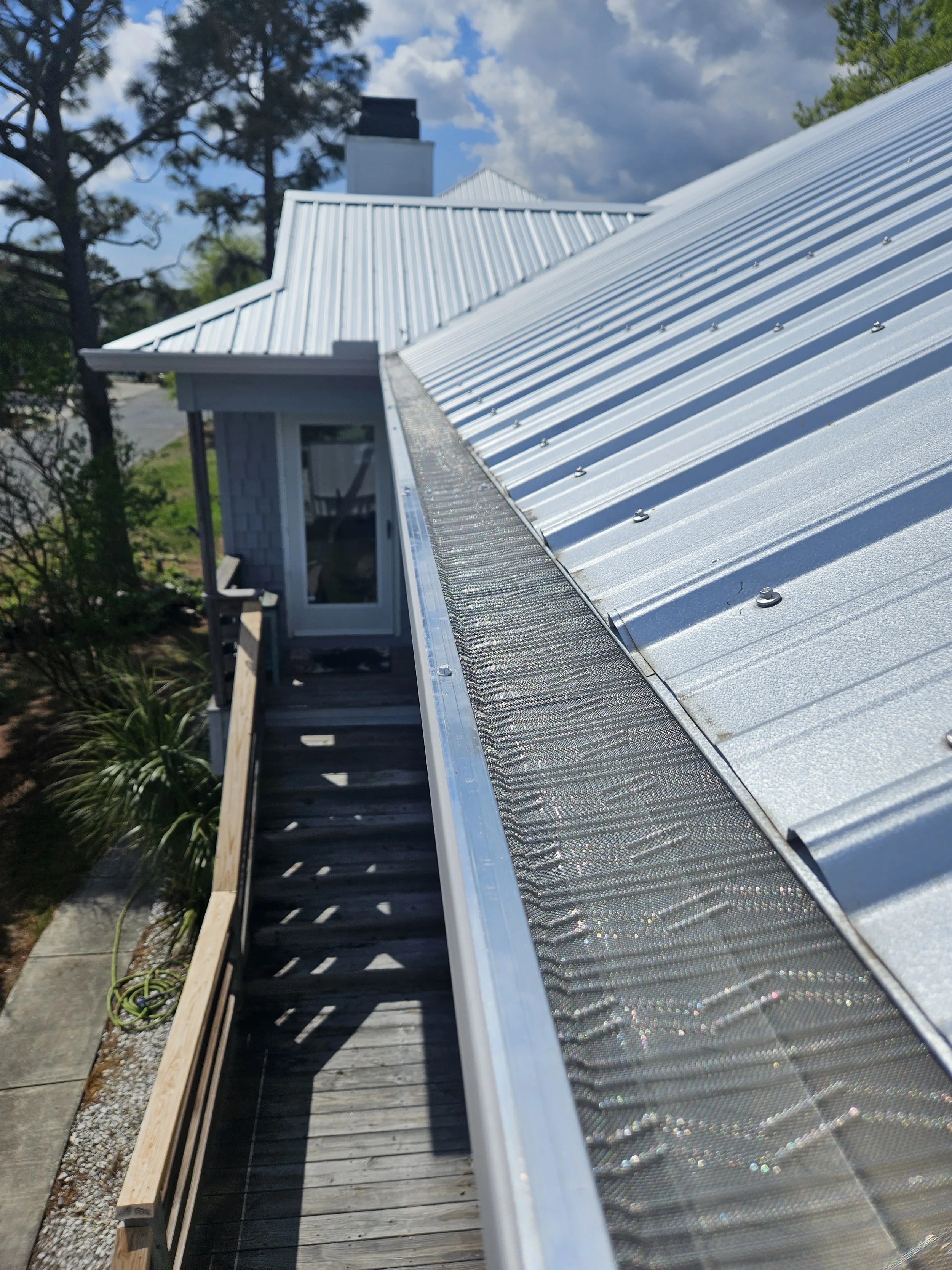 A metal roof on a house with sections under construction, surrounded by trees and a partly cloudy sky.