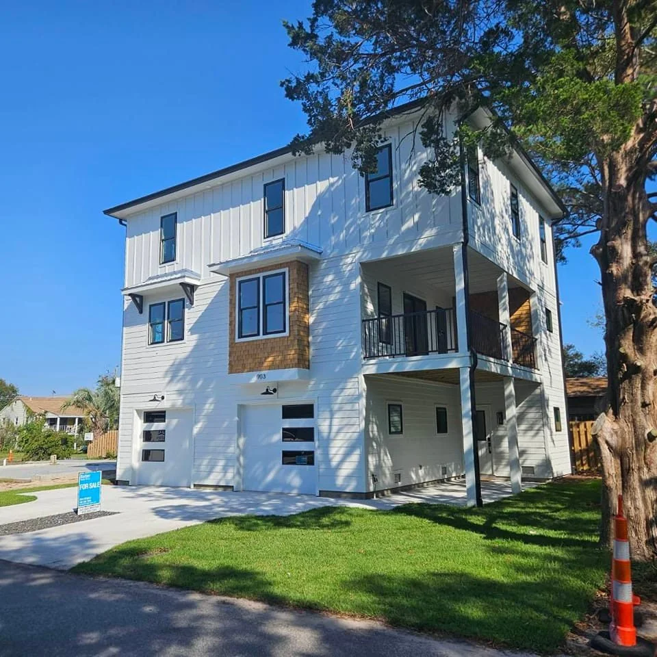 New white multi-story house with garage doors, sitting on a green lawn with a tree nearby, under a clear blue sky.