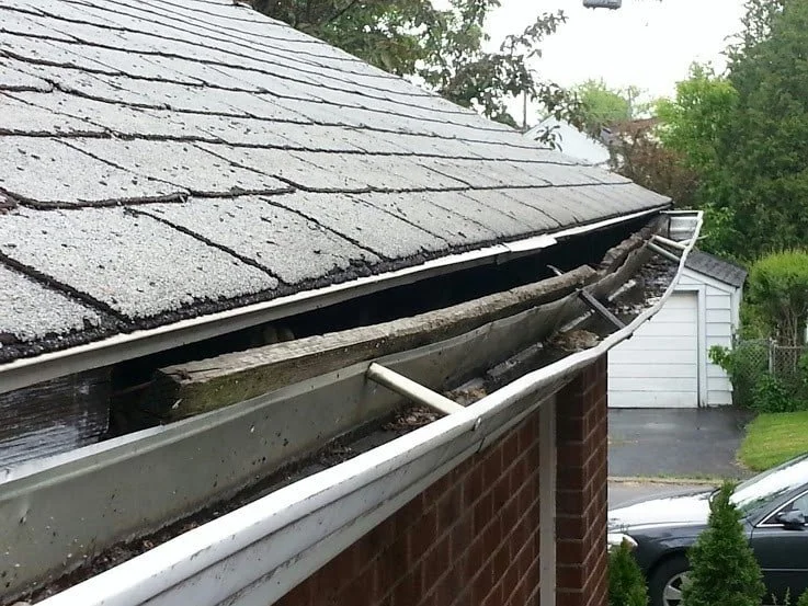 Damaged gutter and roof shingles on a house with exposed wooden support beams.
