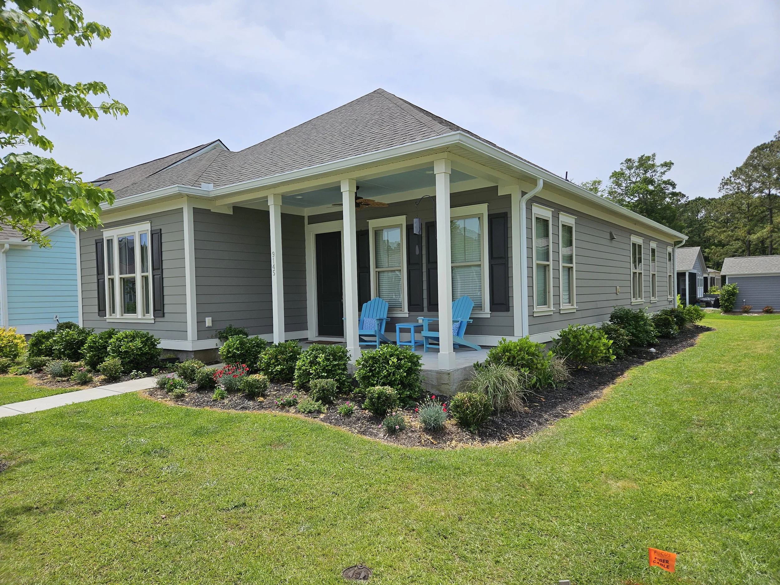 A gray house with black shutters and a small front porch with two blue Adirondack chairs. The house has a shingled roof and white trim. There are bushes and a garden in front of the house, and a large tree to the left. The sky is partly cloudy.
