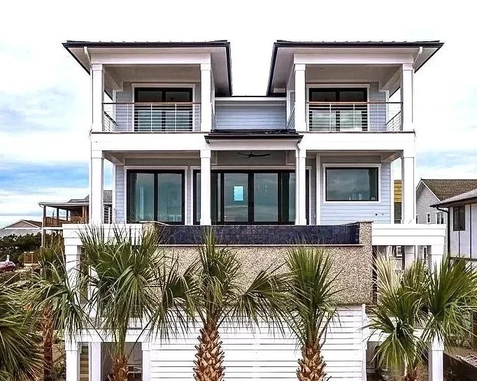 Modern multi-story house with white exterior, large glass sliding doors, and balconies, surrounded by palm trees.