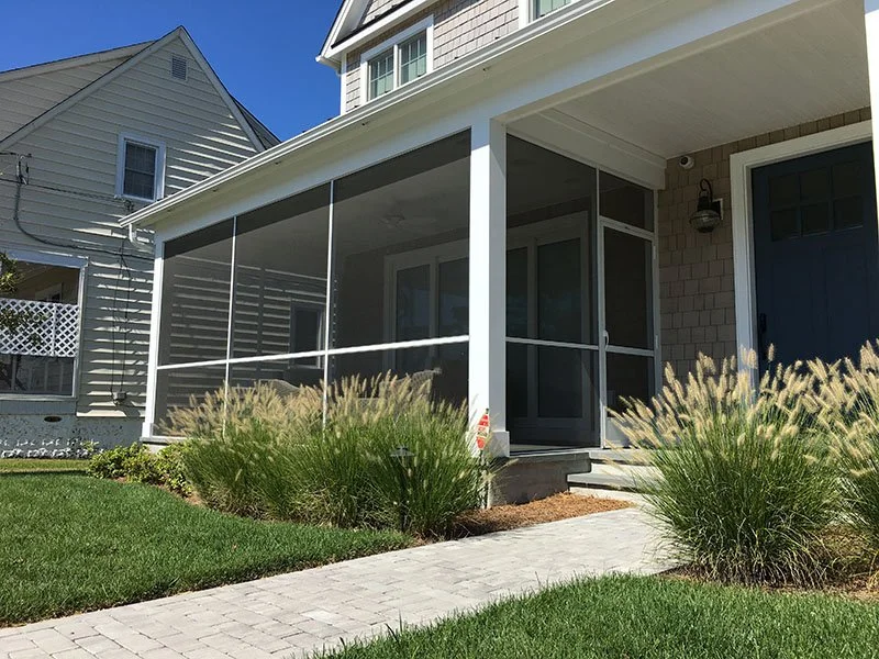 Front view of a house with a screened-in porch, surrounded by green grass and ornamental grasses, with a paved walkway leading to the front door.