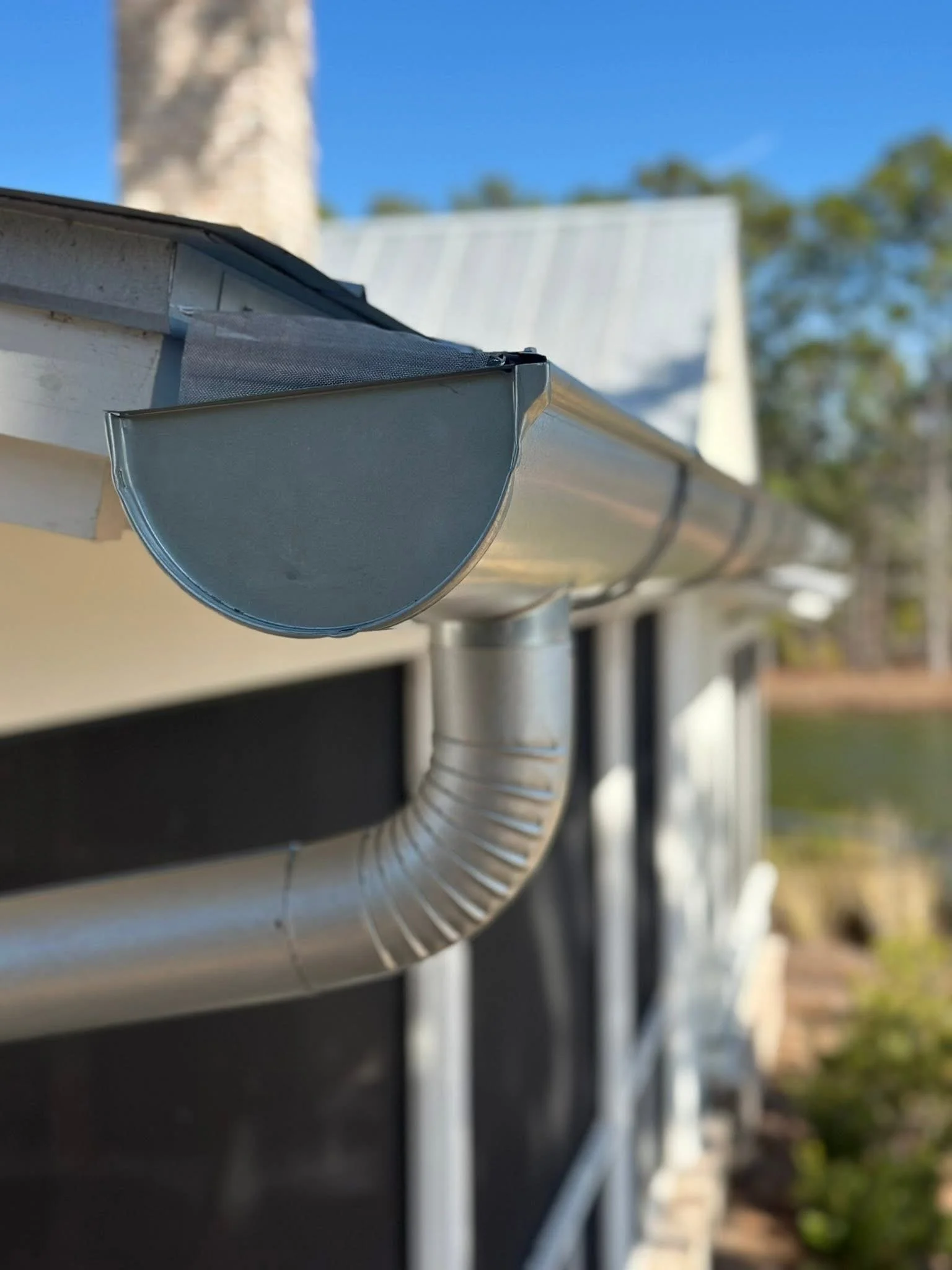Close-up view of a house's rain gutter with a downspout attached, against a background of trees and a blue sky.