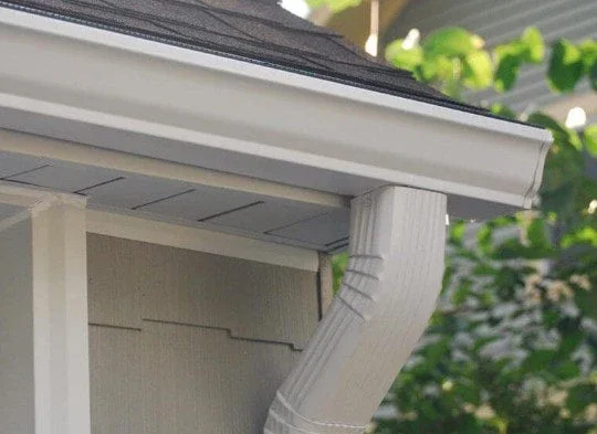 Close-up of the corner of a house's beige siding with white gutter and downspout system against a background of greenery.