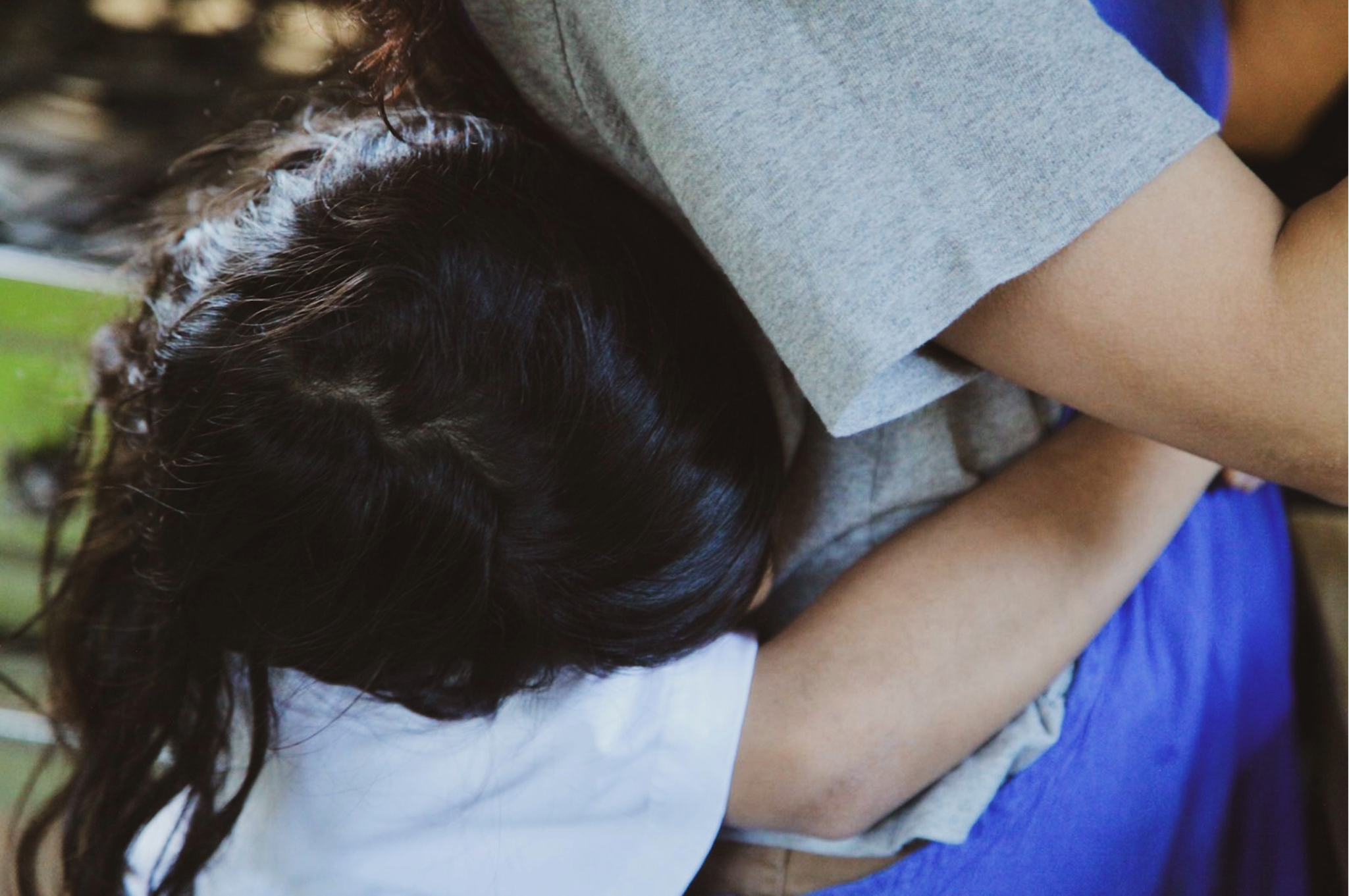 Close-up of a person with dark, curly hair resting their head on their folded arms on a surface, wearing a light gray t-shirt and there is a hint of a blue object in the background.