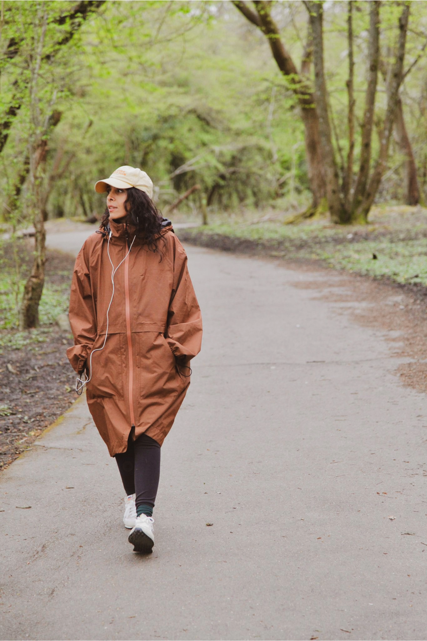 Woman walking on a paved trail in a wooded park, wearing a tan cap, brown rain jacket, black leggings, white sneakers, and listening to earphones.