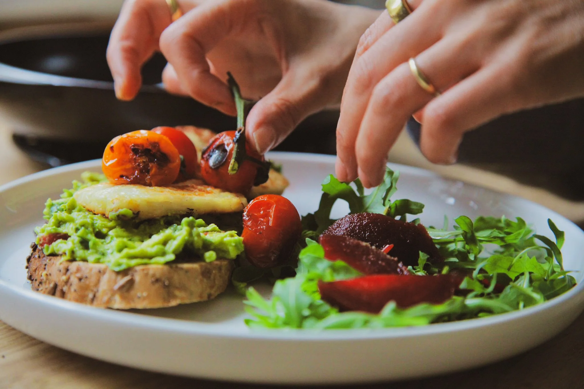Close-up of a plate with avocado toast topped with cherry tomatoes and a fried egg, garnished with greens, and a person's hand placing a cherry tomato on the food.