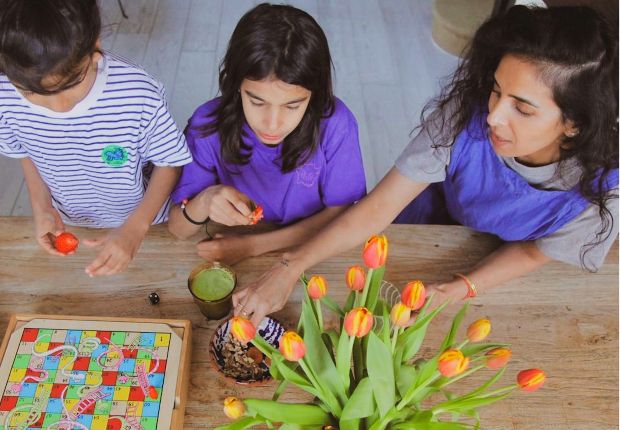 Three children and an adult sitting at a wooden table with a board game, colorful tulips in a vase, and small cups of drinks.