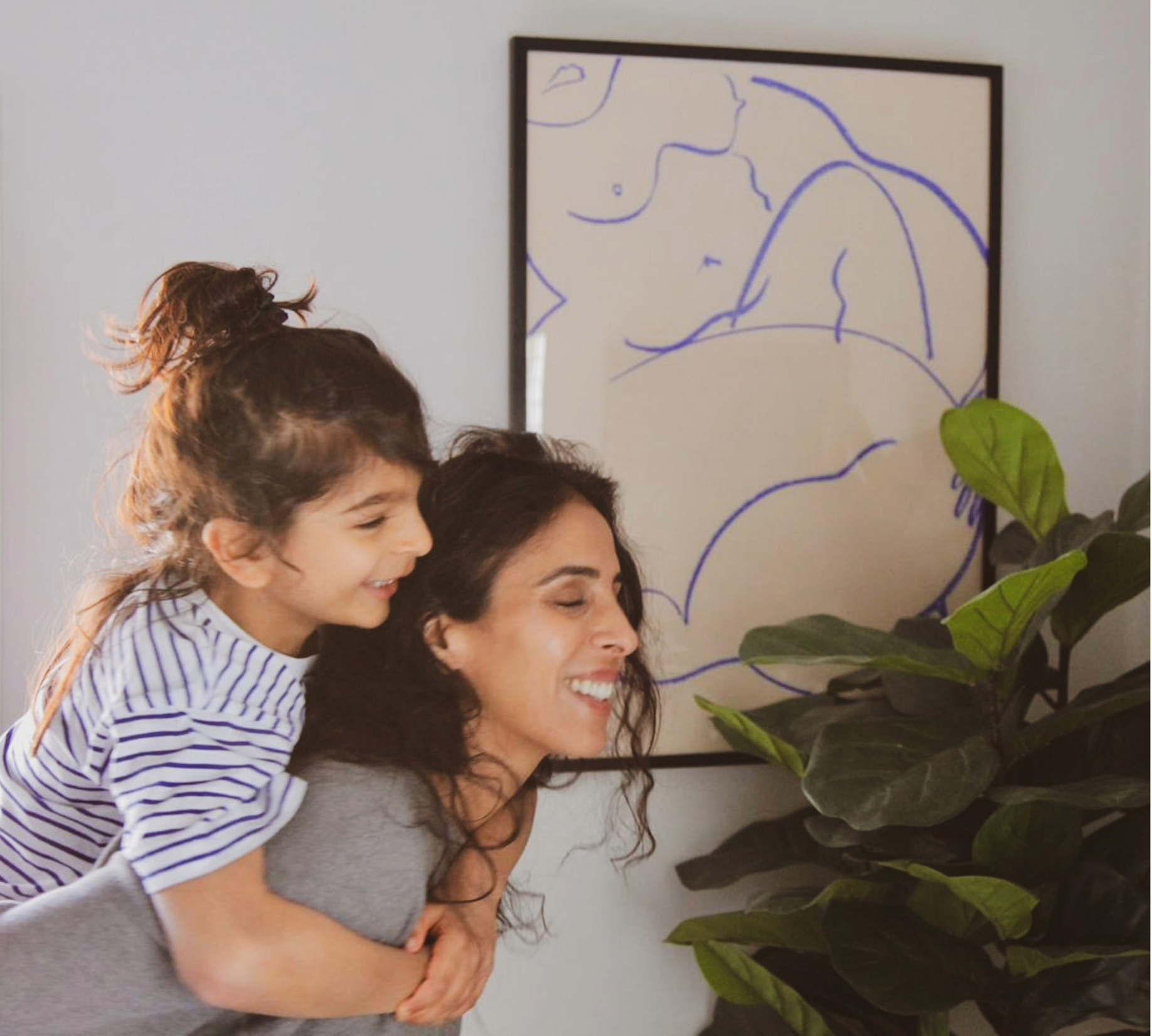 A group of people, including a young girl on an adult woman's back, smiling and laying close to a large green houseplant, with a framed line drawing of a woman on the wall behind them.