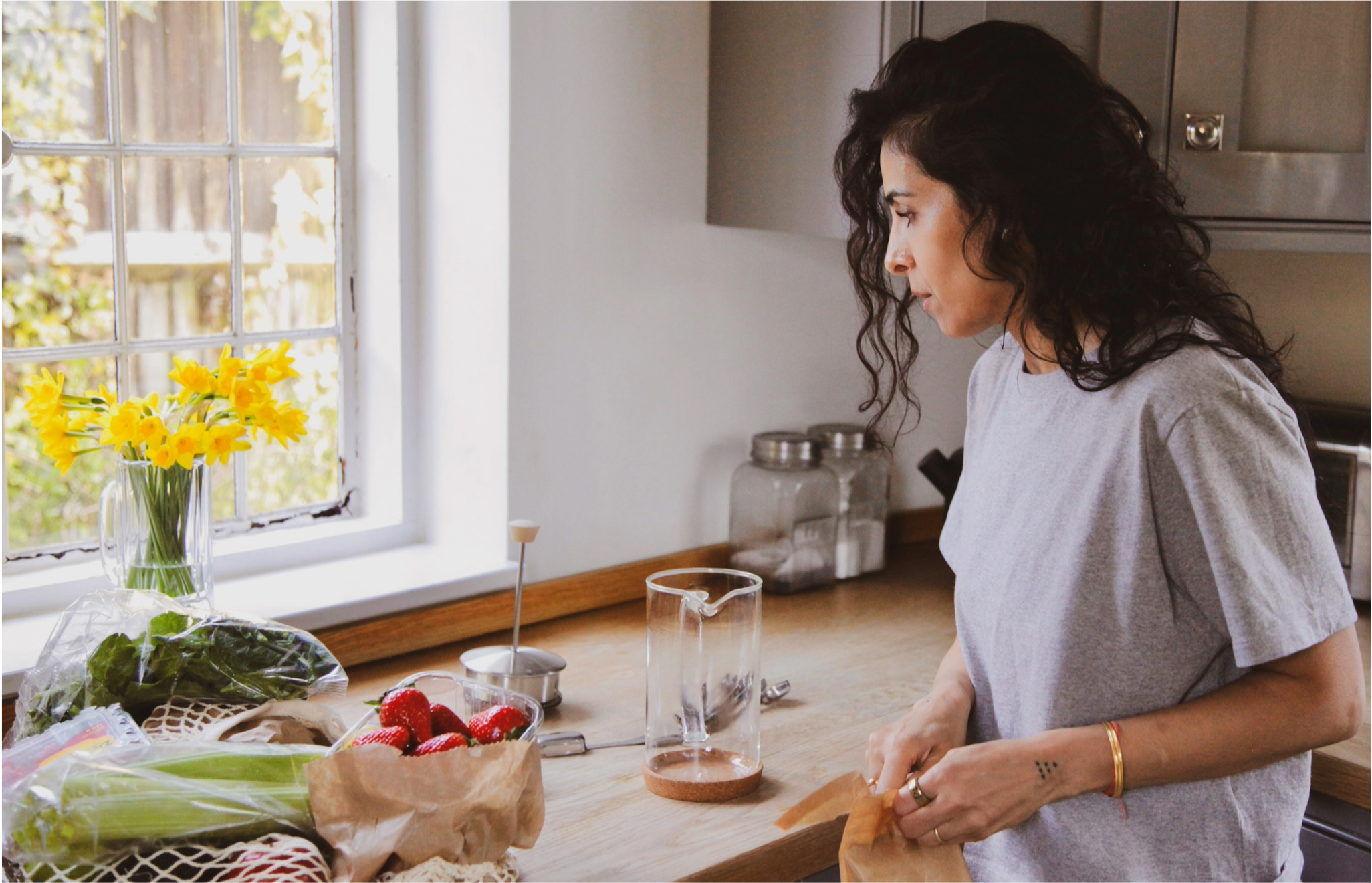 A woman with curly dark hair and tattoos on her right arm is preparing strawberries on a kitchen counter. There are yellow flowers in a glass vase, a plastic bag of vegetables, and strawberries on the counter. She is looking down at what she is doing