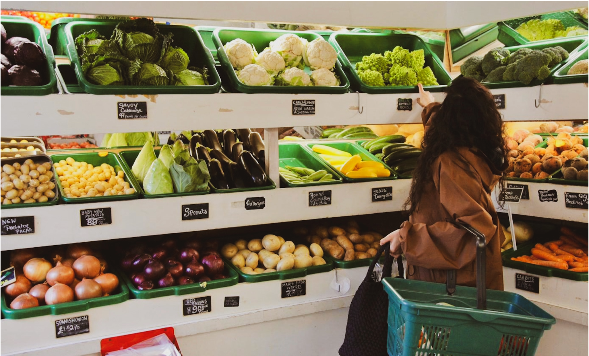 A woman shopping for vegetables at a grocery store, with multiple trays of fresh produce including cauliflower, Romanesco, zucchinis, yellow squash, eggplants, potatoes, onions, carrots, and other vegetables on display.