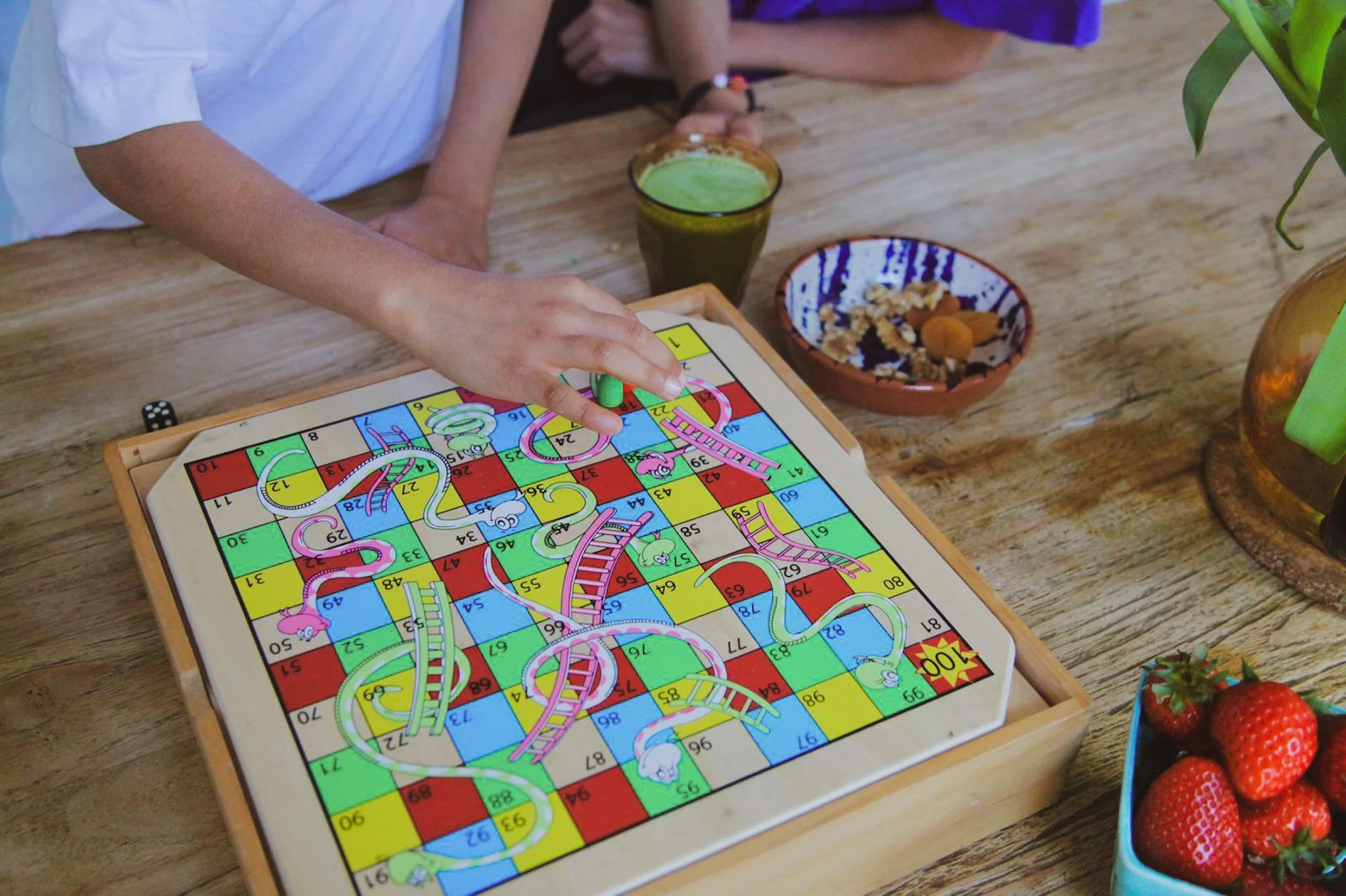 A colorful Snakes and Ladders board game on a wooden table with various snacks and drinks around it.