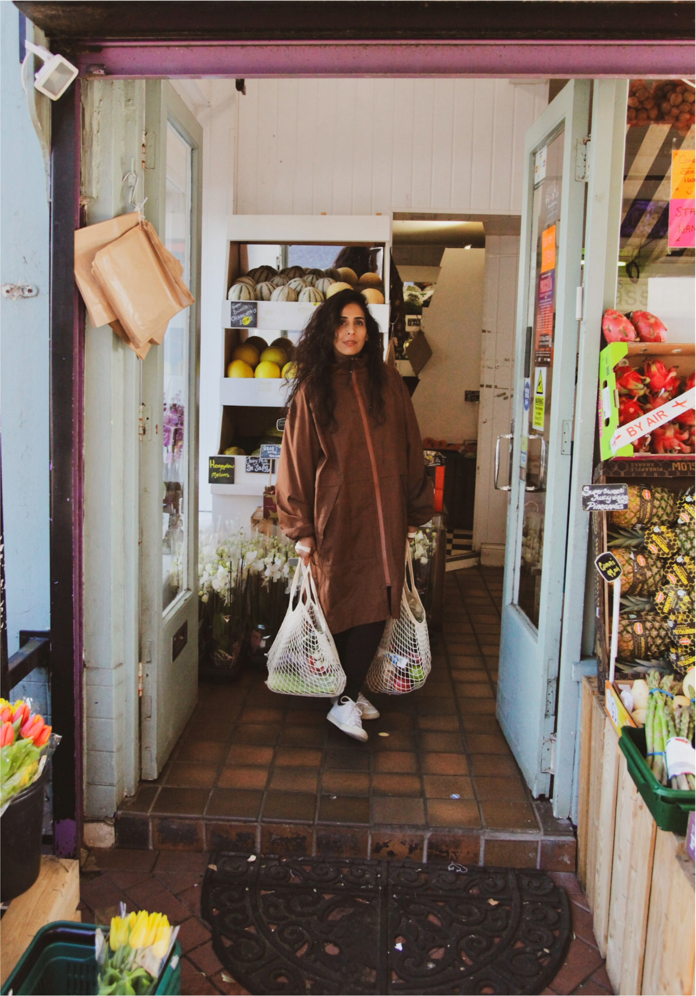 A woman stands inside a small grocery store, carrying two plastic shopping bags filled with groceries. The store has shelves with fruits, vegetables, and flowers, with a rustic tiled floor and a black decorative welcome mat at the entrance.