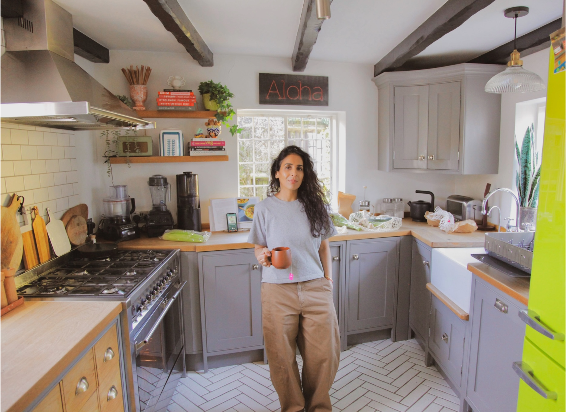 Woman with curly dark hair holding a mug in a kitchen with gray cabinets, a stove, open shelves, and a white subway tile backsplash. She is wearing a gray t-shirt and tan pants.