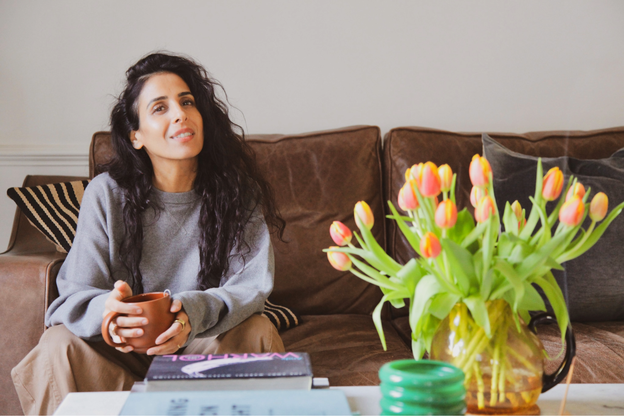 A woman with long dark curly hair sitting on a sofa holding a coffee mug, with a large bouquet of pink and yellow tulips on the table in front of her.