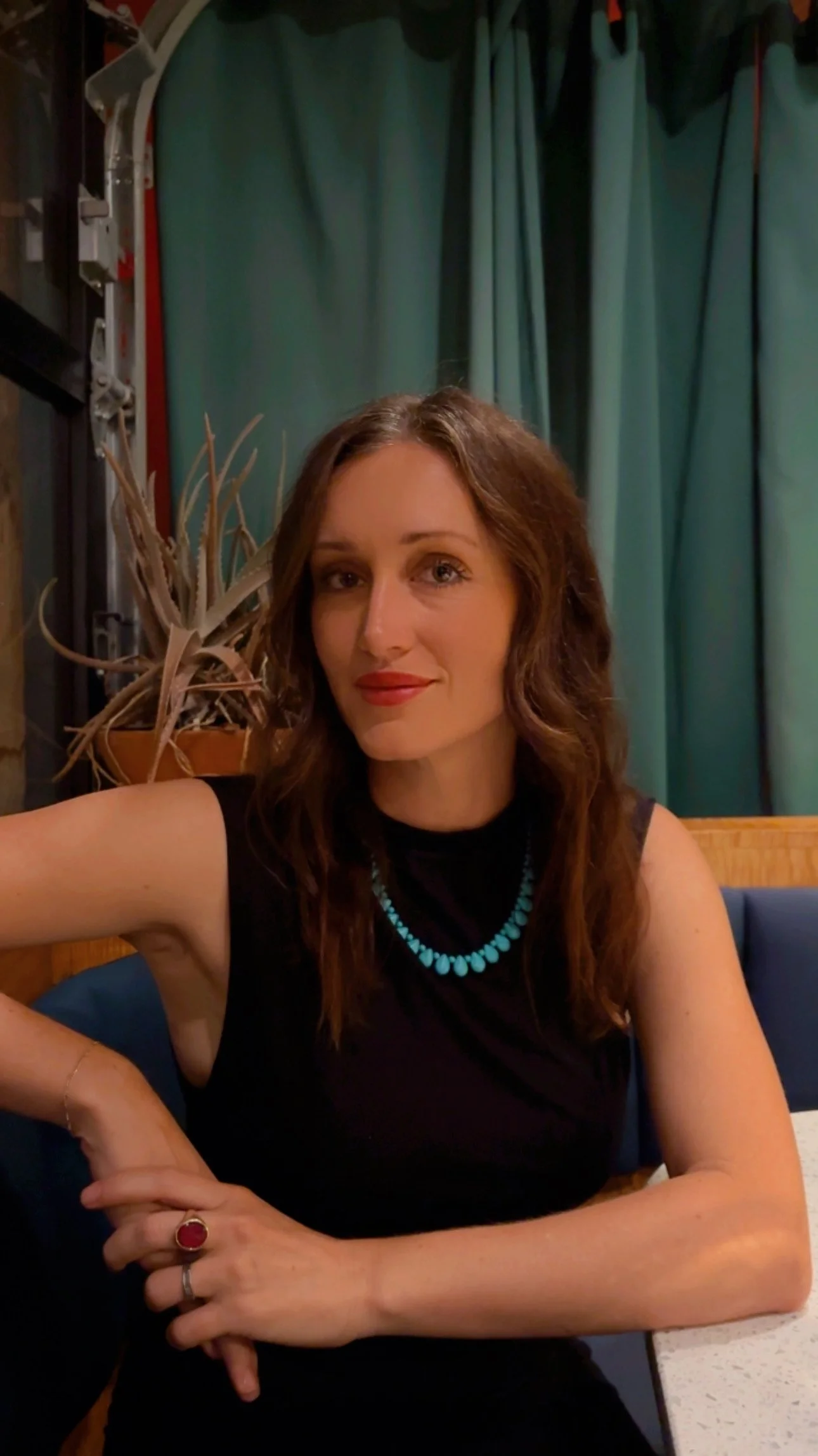 A woman with wavy brown hair sitting at a table in a restaurant, wearing a black sleeveless top, turquoise necklace, a ring with a red gemstone, and a delicate bracelet, with a green curtain and potted plant in the background.