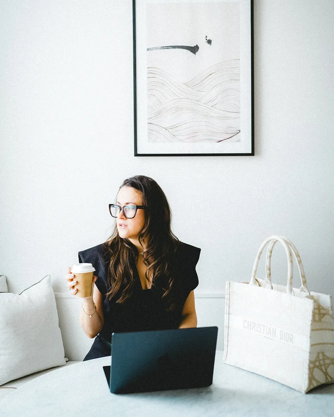 A woman with glasses and dark hair sitting at a white table holding a coffee cup. There is a laptop in front of her and a large Christian Dior tote bag on the table. A framed abstract black and white artwork hangs on the white wall behind her.