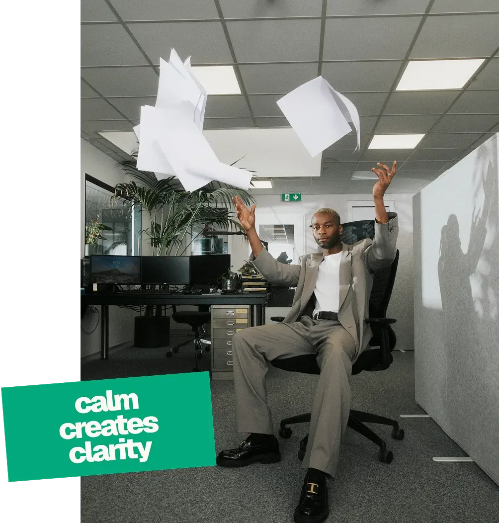A man in a suit sitting in an office chair with papers flying around him, and a green sign in the corner that reads 'calm creates clarity'.