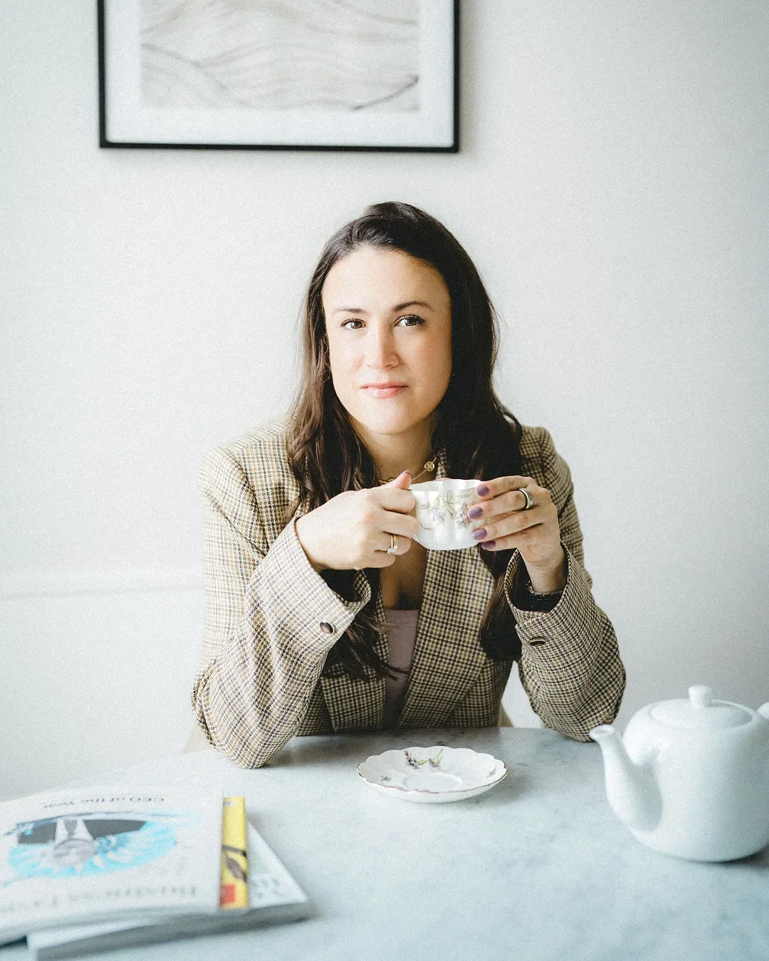 A woman with dark hair wearing a checkered blazer sitting at a table, holding a teacup and looking at the camera. There is a teapot and a magazine on the table.