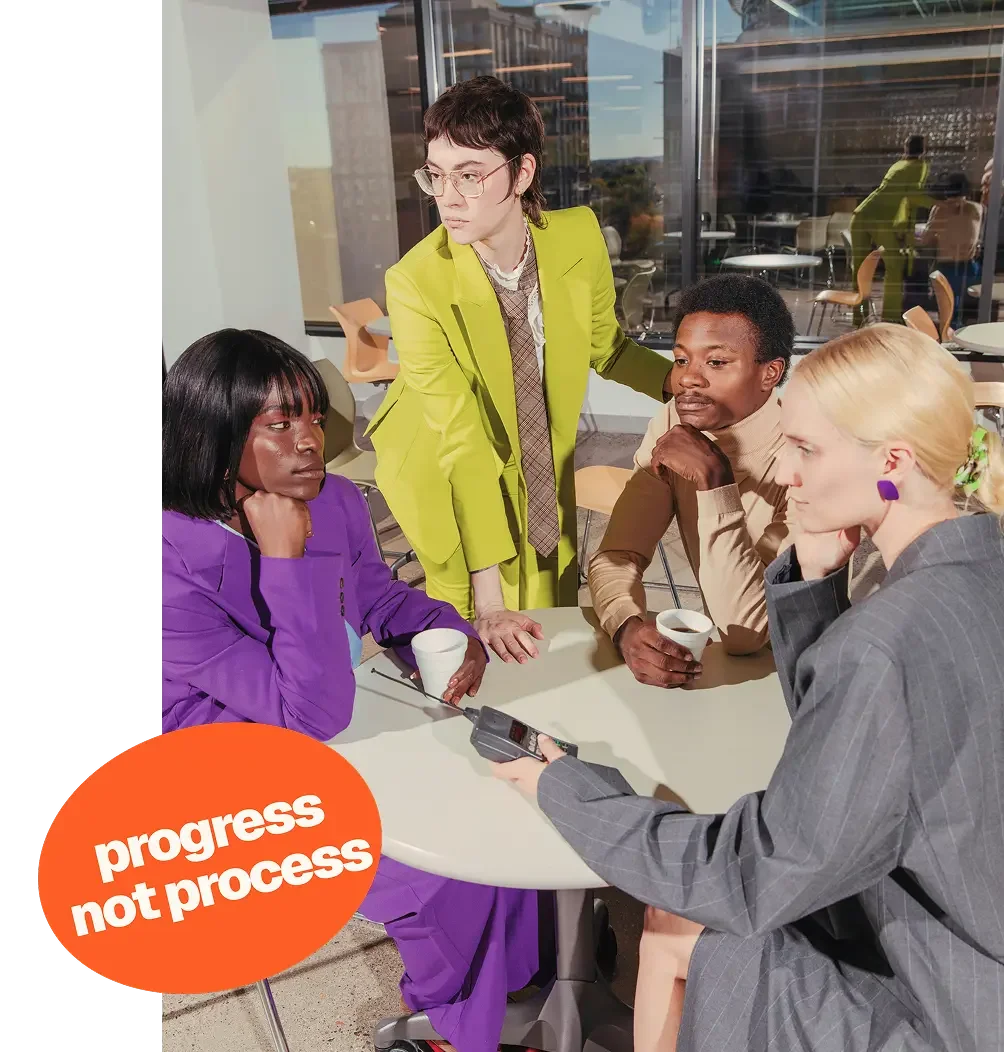Group of four women engaged in discussion at a café table, with one woman holding a recording device, inside a modern glass-walled building during daytime. An orange sticker on the image reads 'progress not process.'