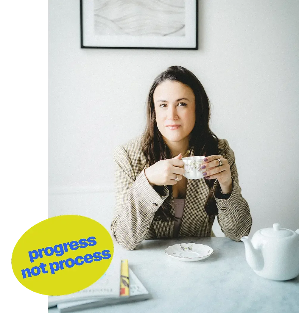 A woman with dark hair, wearing a plaid blazer, sitting at a table with a teacup in her hands. There is a teapot, a small plate, and a magazine or book on the table. A framed picture hangs on the plain white wall behind her. A yellow speech bubble with blue text reads 'progress not process.'
