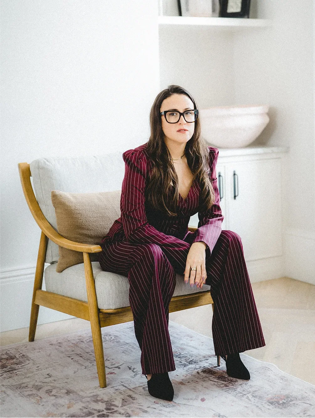 Carly Jenner, a woman with long dark hair, wearing glasses, a maroon pinstriped suit, sitting on a beige armchair in a room with a white wall and a rug.