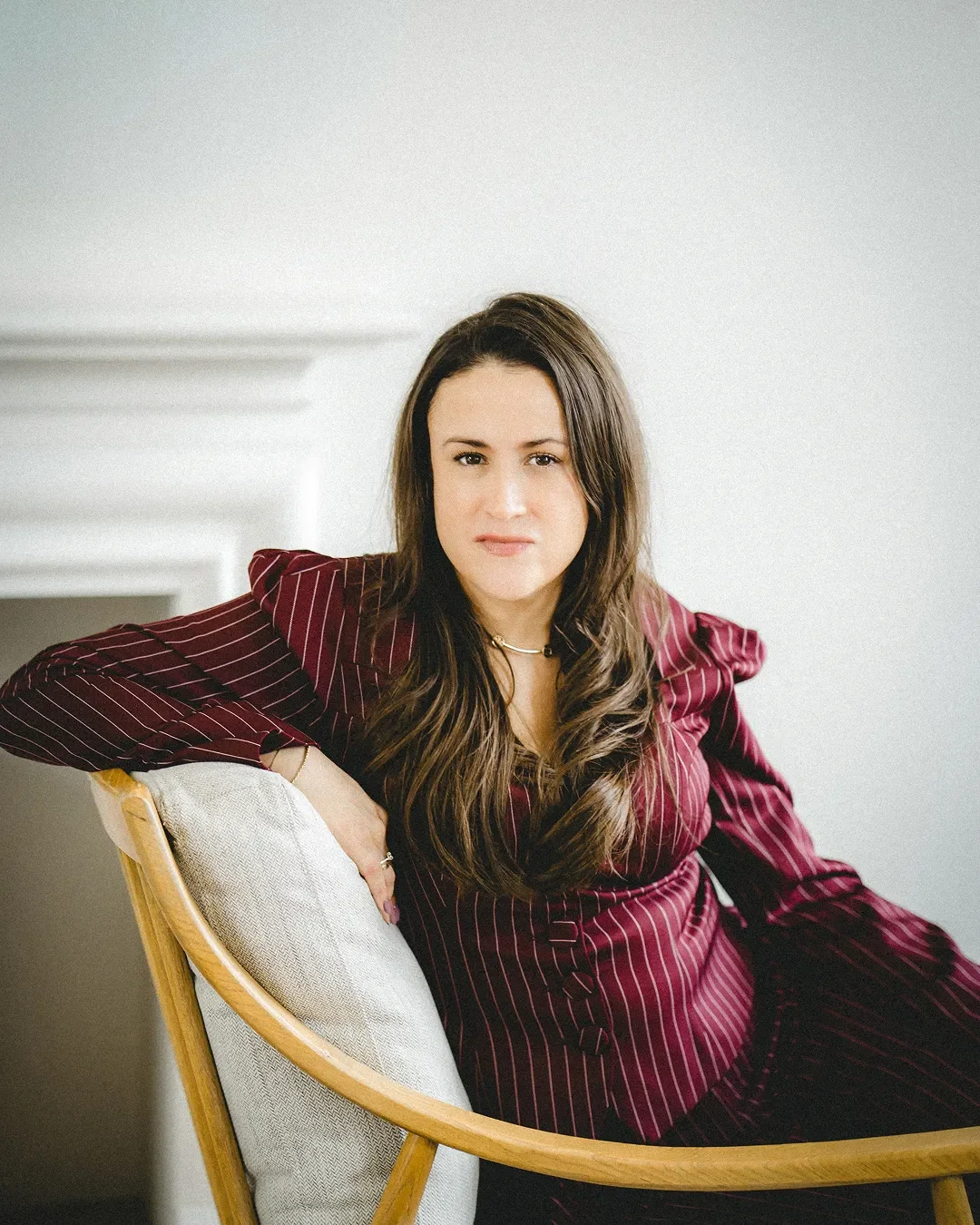A photo of Carly Jenner, a woman with long brown hair sitting on a light-colored chair with a wooden frame, wearing a burgundy pinstripe dress, against a light-colored wall.