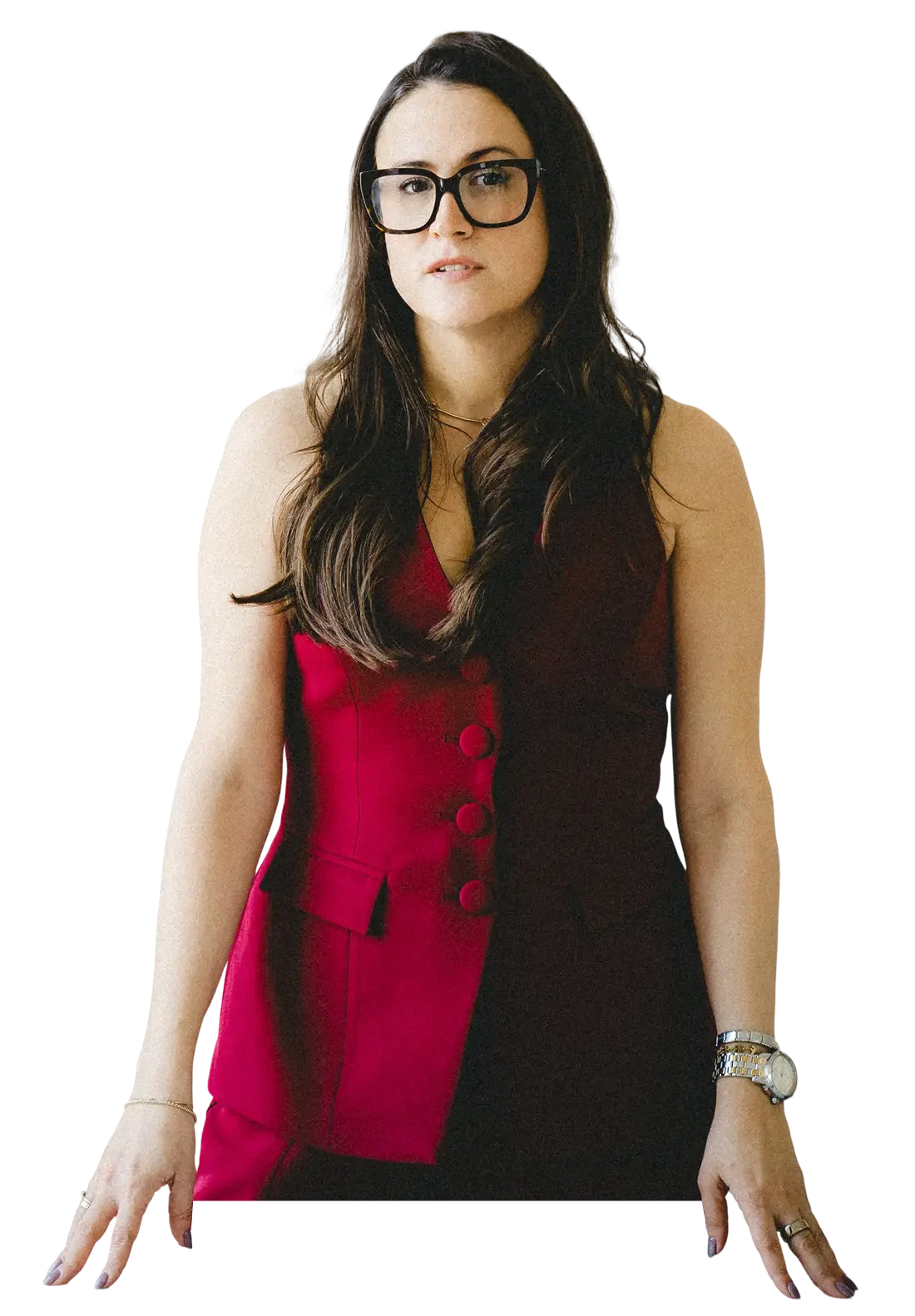 A photo of Carly Jenner, a woman with long brown hair, wearing glasses, a red and black dress with large buttons, and a silver watch, standing with her hands on a surface.