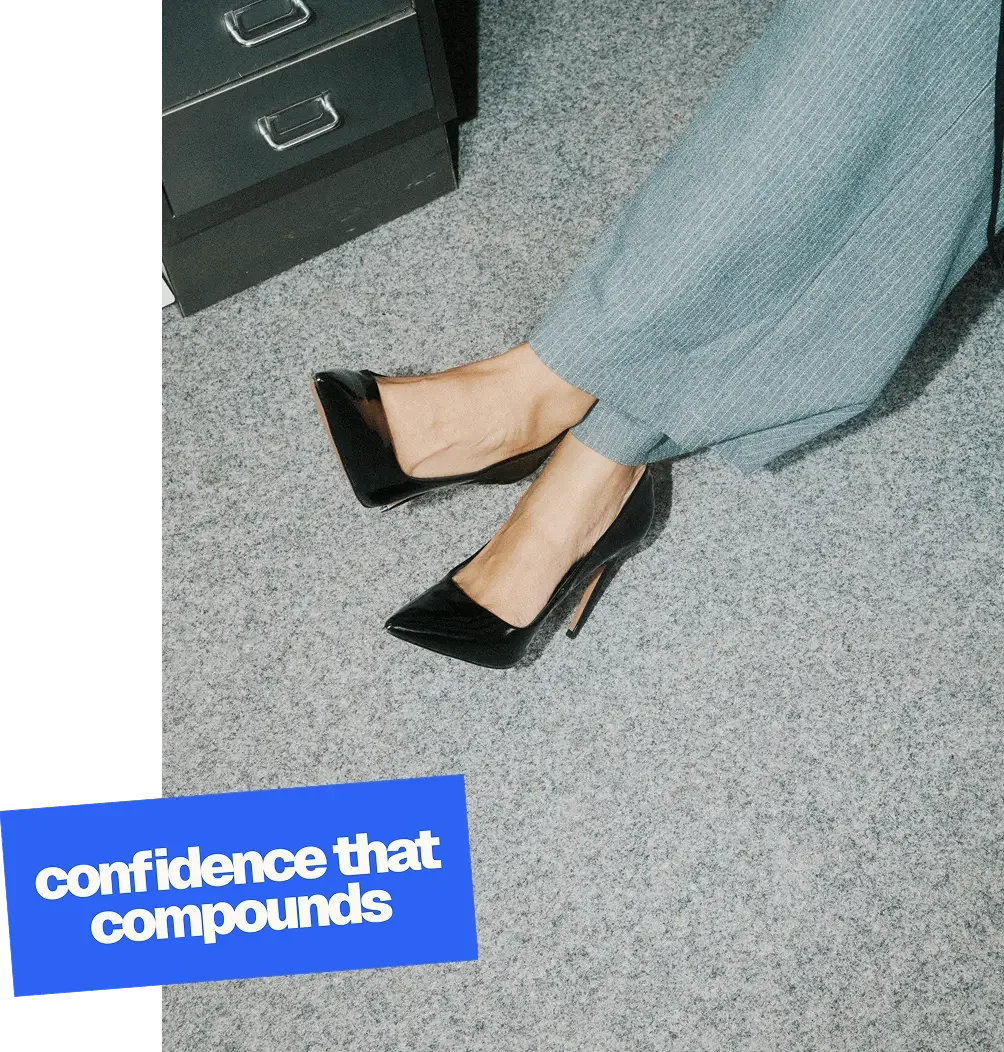 Close-up of a person sitting on the floor wearing gray pinstripe pants and black high-heeled shoes, with a blue text box that reads 'confidence that compounds'.