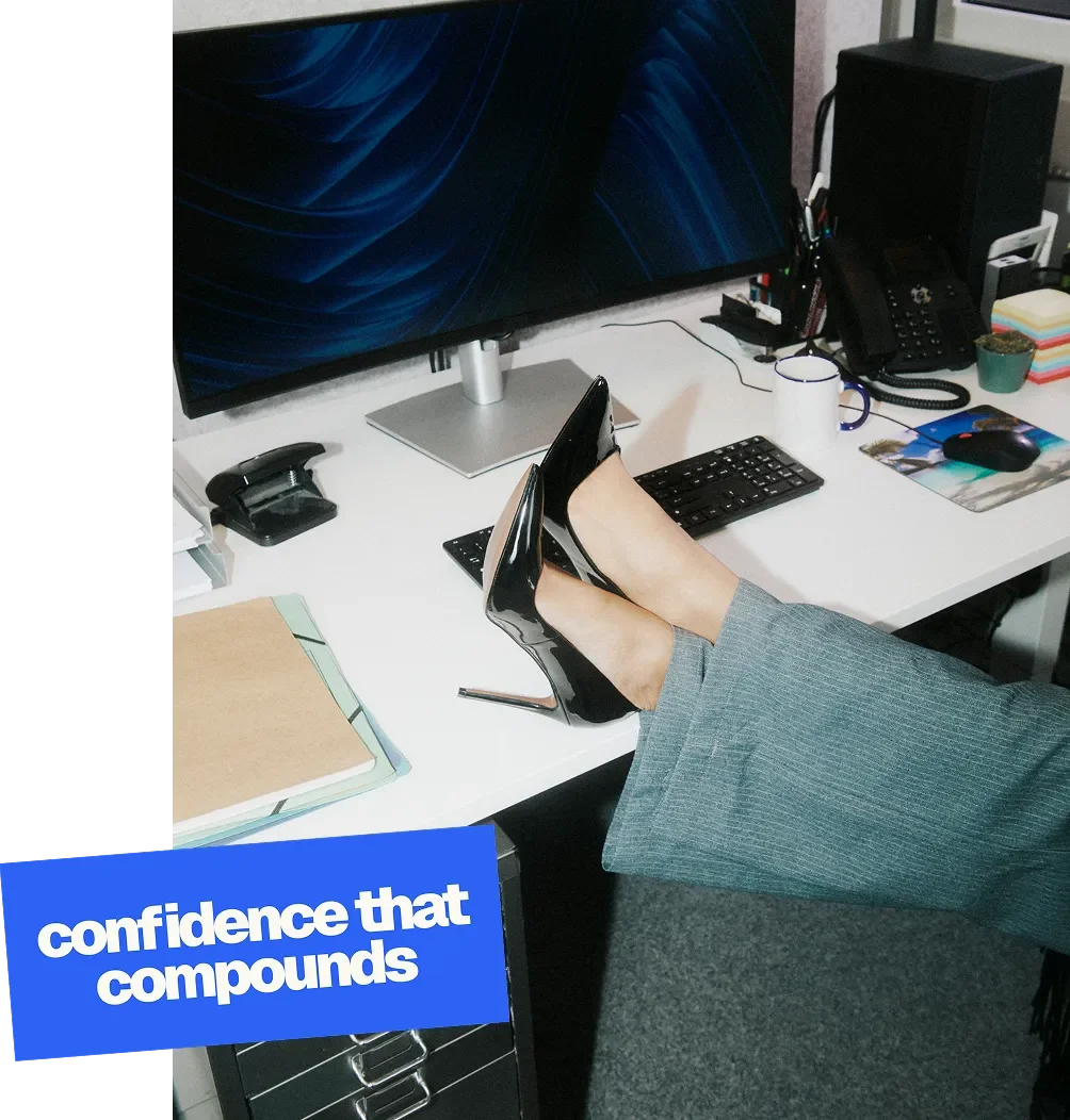 A person wearing gray office pants and black high heels with their feet resting on a cluttered white office desk. The desk has a monitor, keyboard, mouse, a cup, a telephone, a pot with a plant, sticky notes, and a folder. There is a blue sticky note on a file cabinet with the text "confidence that compounds."