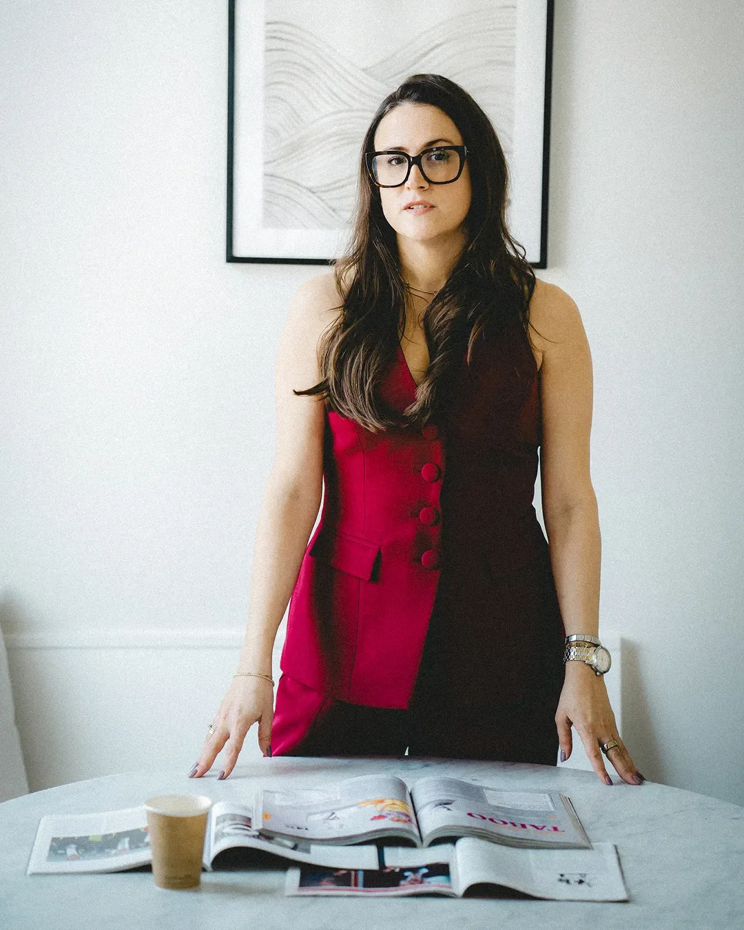 A photo of Carly Jenner, a woman with long dark hair, wearing glasses and a sleeveless dress with a red and black color scheme, standing behind a table with magazines and an empty paper cup, in a modern room with abstract wall art.