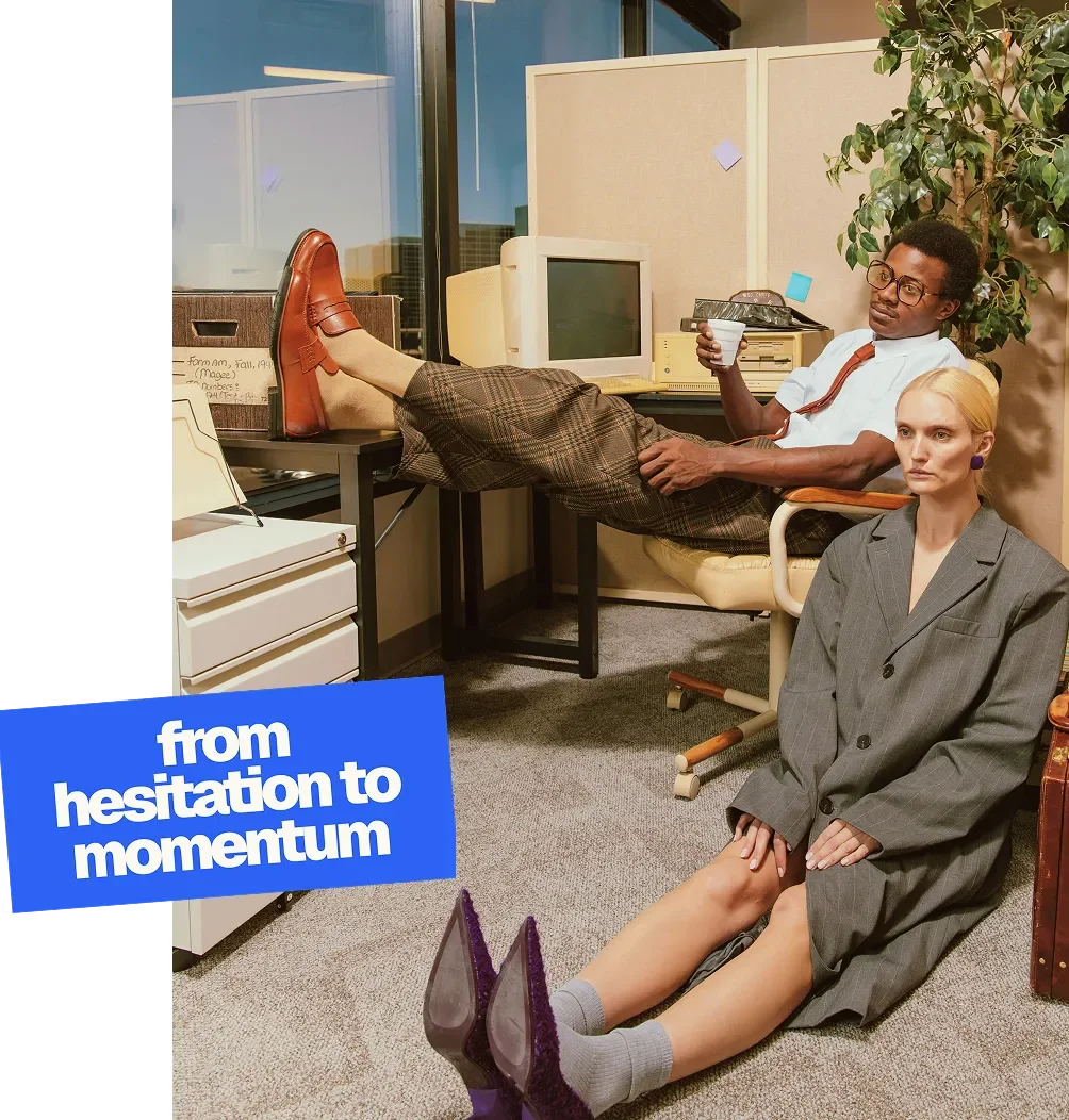 Two people in an office, one man relaxing in a chair with feet up on the desk holding a cup, and a woman sitting on the floor with legs extended and head straight, both dressed in business attire. There's a label that reads 'from hesitation to momentum'.