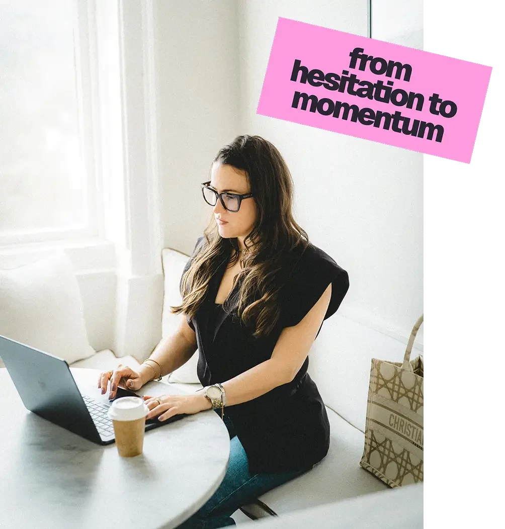 A woman with long dark hair and glasses working on a laptop at a table, with a coffee cup and a bag nearby, and a sign on the wall that says 'from hesitation to momentum'.